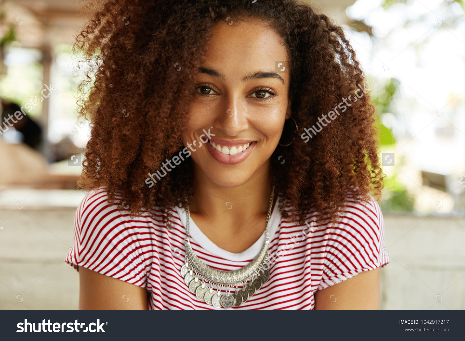 Portrait of happy of dark skinned female with curly bushy Afro hairstyle, smiles broadly into camera, rests in cafe with boyfriennd, being glad to have much spare time. People, ethnicity concept #1042917217