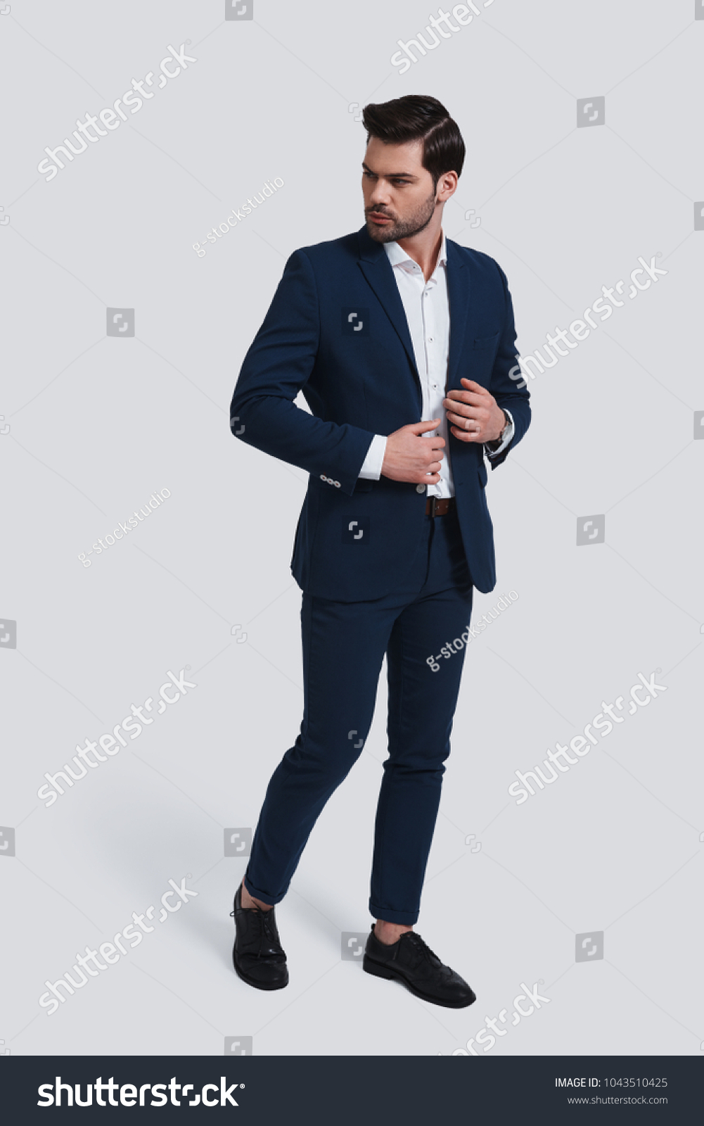 Used to look perfect. Full length of handsome young man in full suit looking away and adjusting his jacket while standing against grey background #1043510425