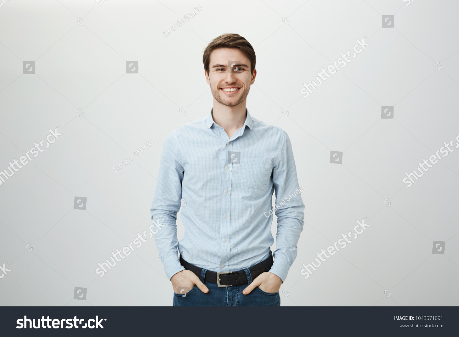Confidence and business concept. Portrait of charming successful young entrepreneur in blue-collar shirt, smiling broadly with self-assured expression while holding hands in pockets over gray wall #1043571091