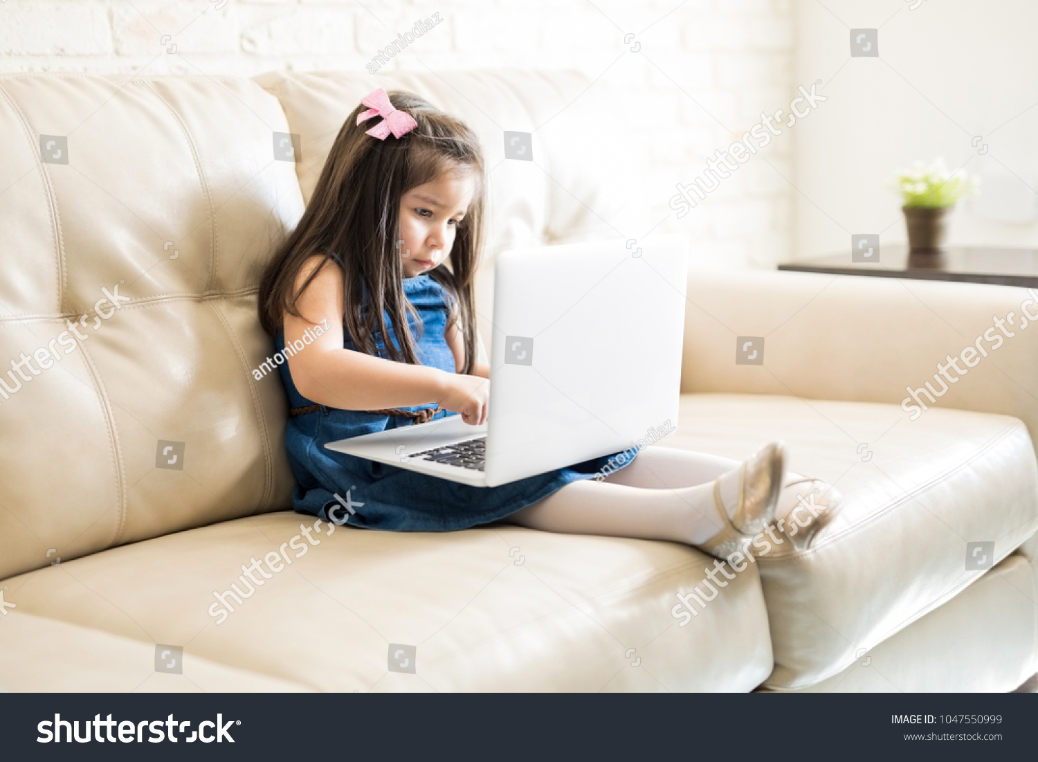 Little hispanic girl playing games on laptop computer while sitting on sofa at home #1047550999