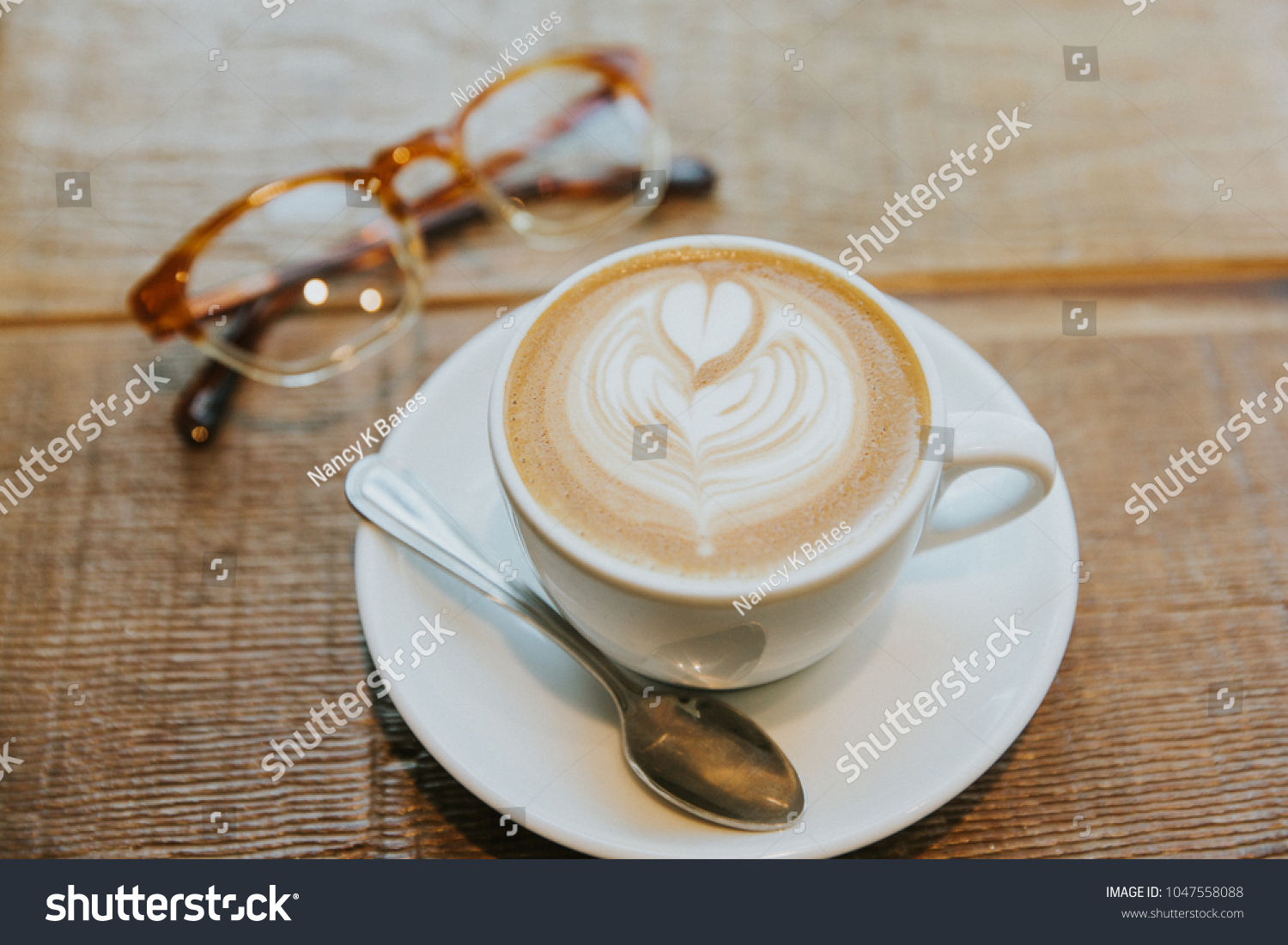 Latte art heart in simple white mug with plate and spoon on a wooden table with a pair of eye glasses #1047558088