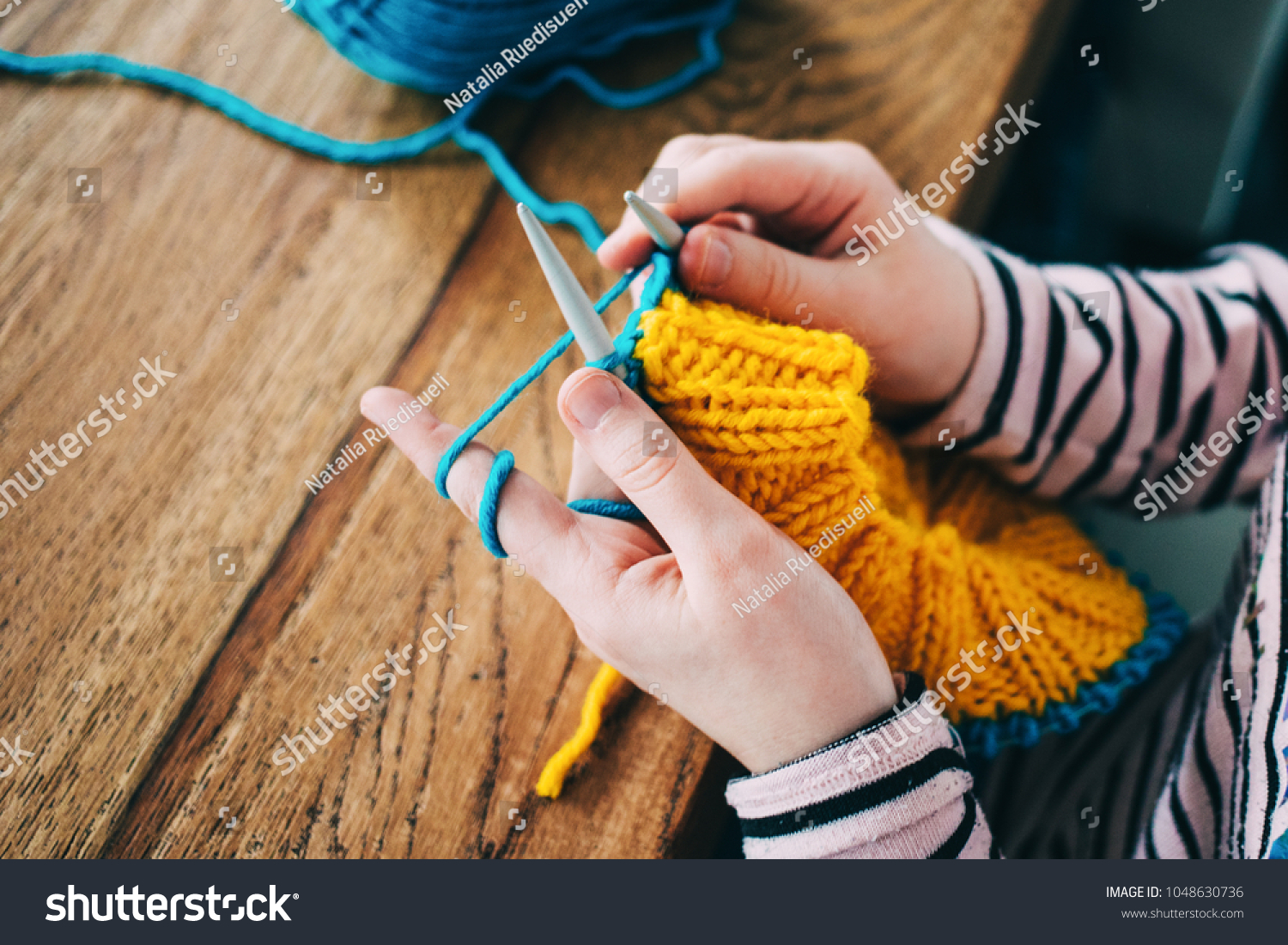Young girl knitting a circle scarf with yellow and blue coloured yarn. Sitting at the wooden table, close up of the knitting needles. #1048630736