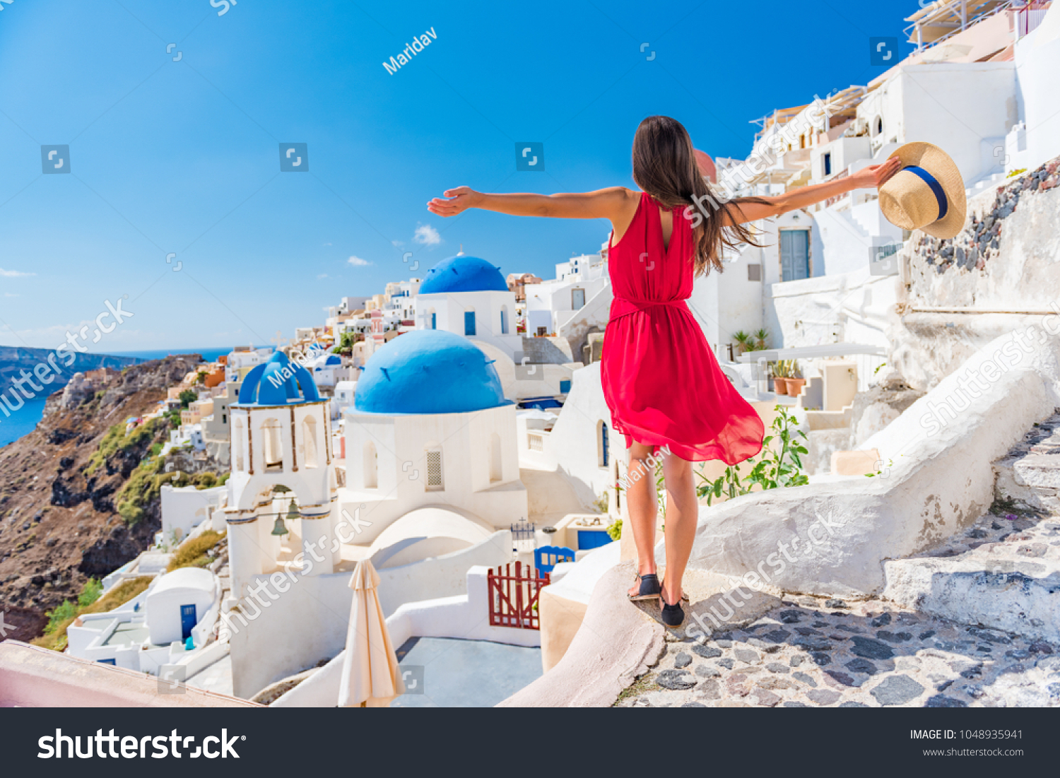 Europe travel vacation fun summer woman dancing in freedom with arms up happy in Oia, Santorini, Greece island. Carefree girl tourist in European destination wearing red fashion dress. #1048935941