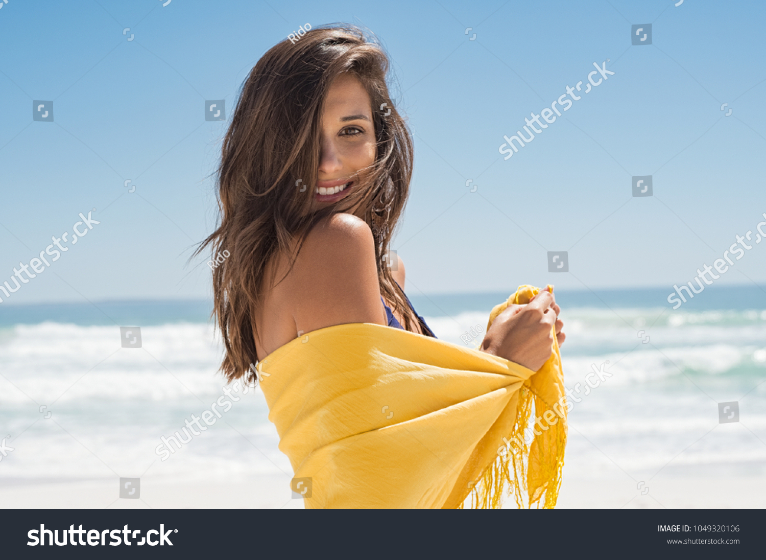 Cheerful young woman in yellow sarong at beach. Happy smiling girl enjoying the beach and looking at camera. Latin tanned woman feeling refreshed in yellow scarf during summer vacation. #1049320106
