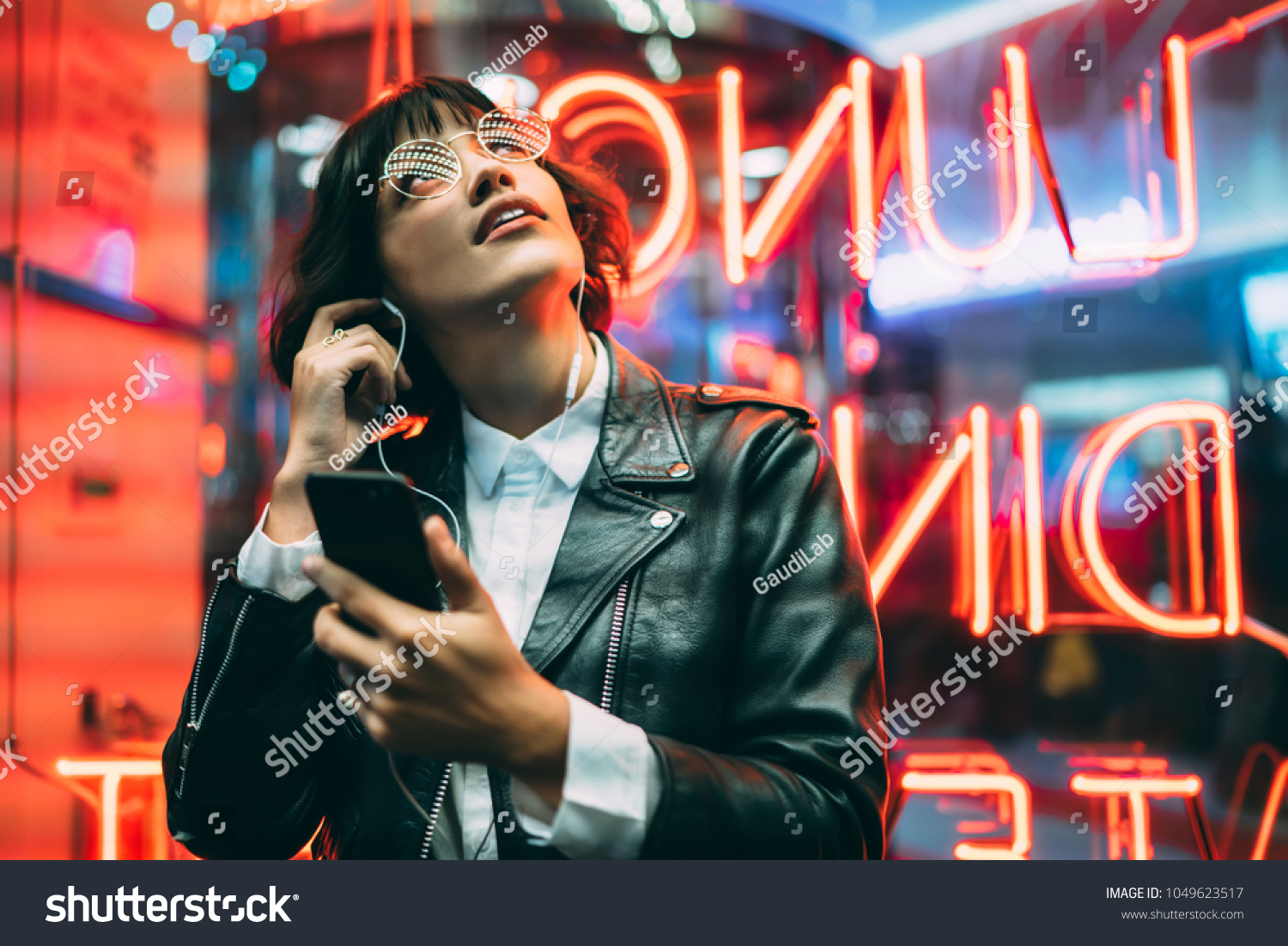 Excited fashion female lover of music dressed in stylish leather jacket listening songs online in earphones connected to smartphone while enjoying night lights and neon illumination in New York City #1049623517