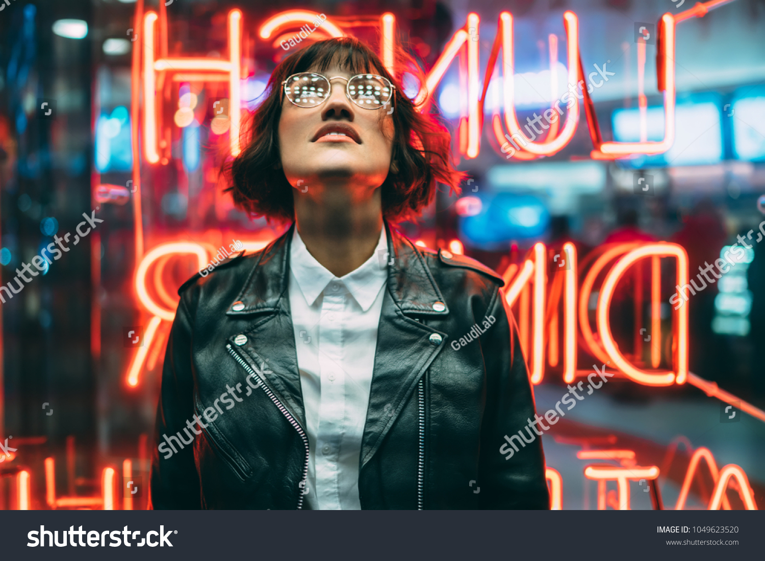 Stylish brunette woman in trendy apparel and eyewear looking up enjoying nightlife in city. Gorgeous fashion hipster girl in leather jacket standing outdoors on street with neon city illumination #1049623520