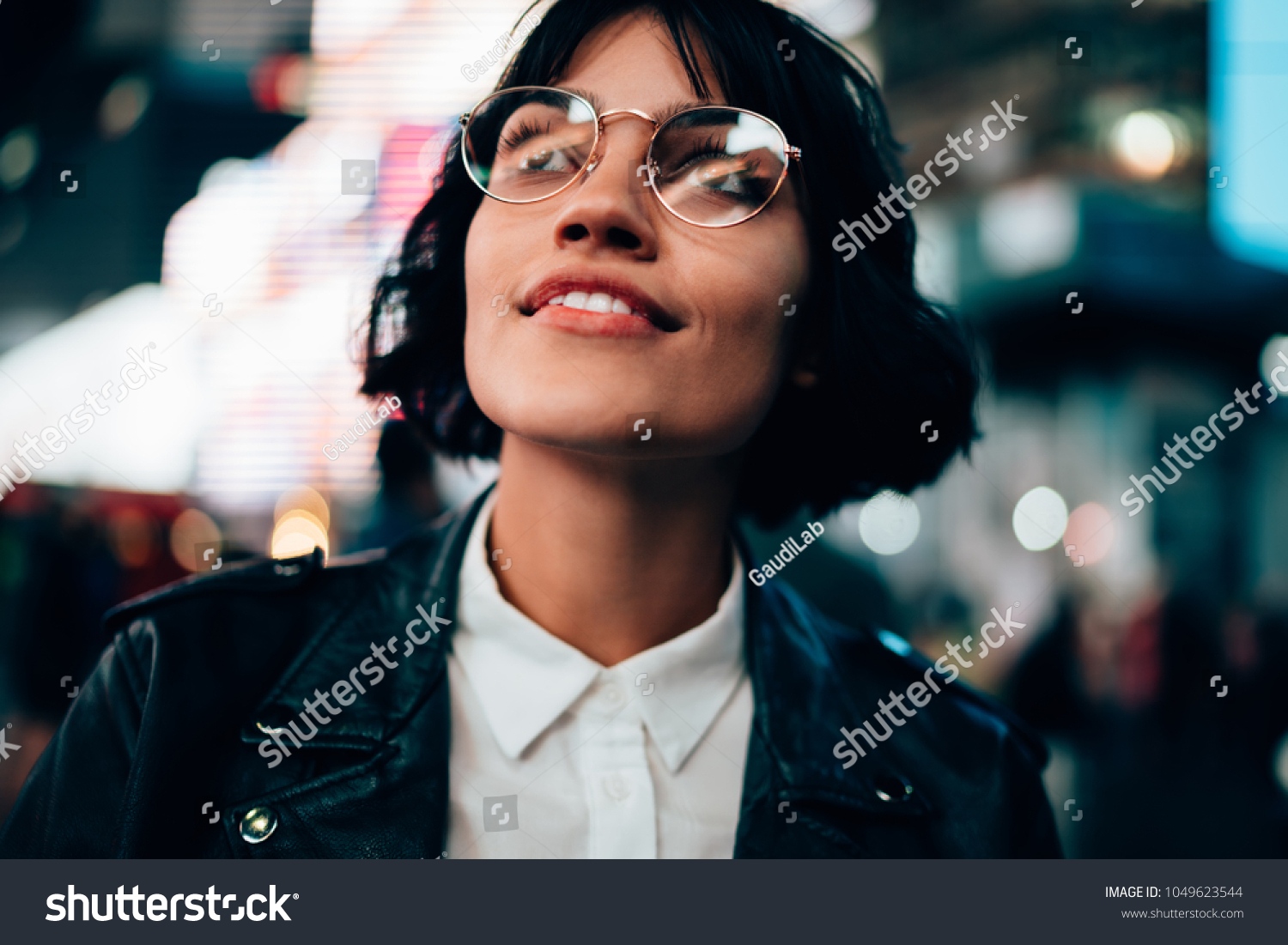 Fascinated pretty hipster girl in stylish eyeglasses amazed with night New York city walk in urban setting. Closeup excited stylish young woman standing in night city against blurred bokeh lights #1049623544
