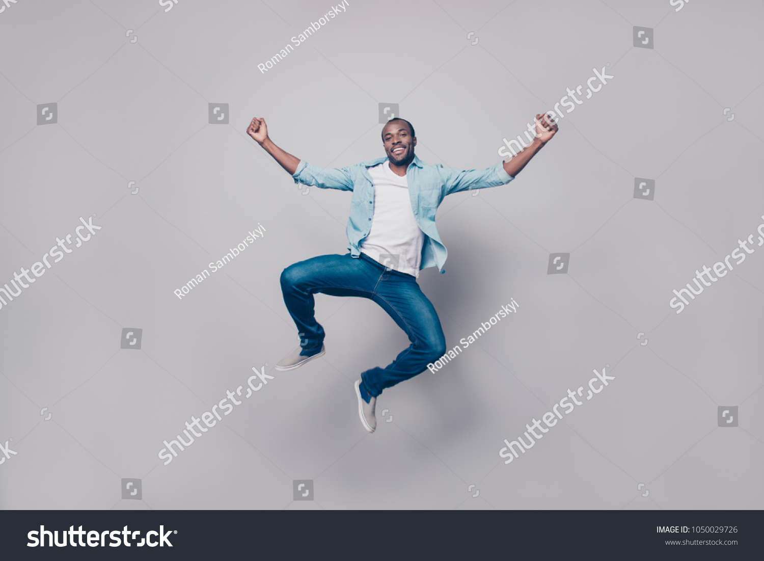 Full length, size portrait of confident attractive, joyful, cheerful, positive, glad guy in shirt, sneakers jumping with raised fists, looking at camera, isolated on grey background #1050029726