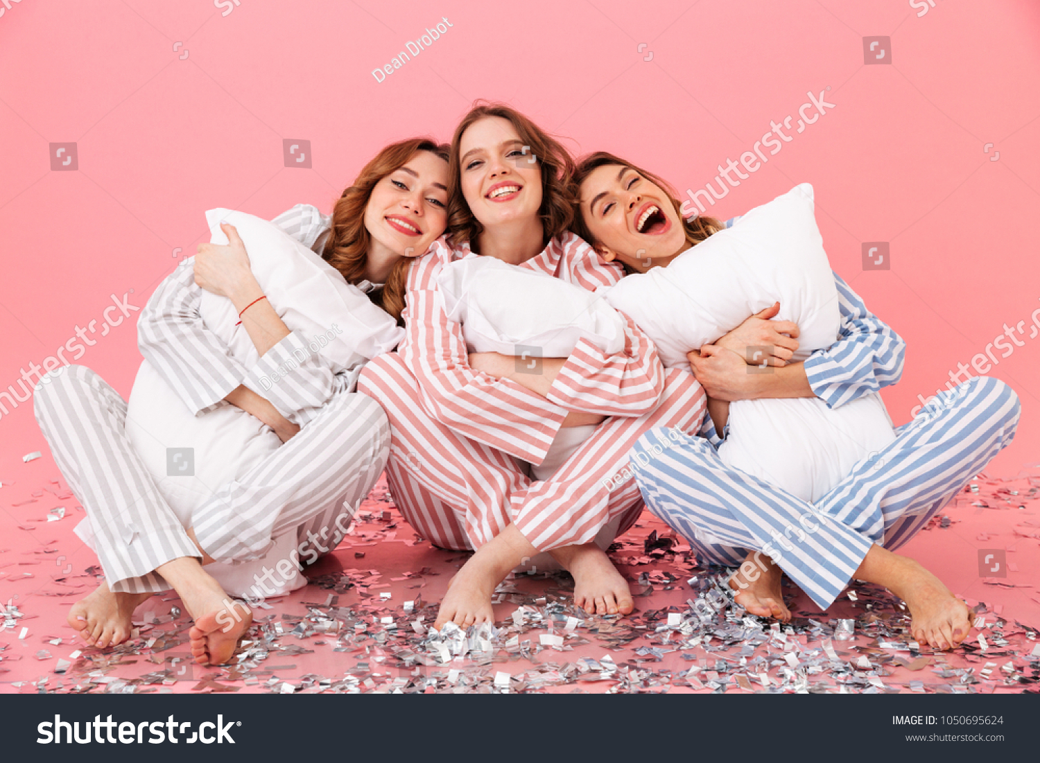 Attractive women 20s wearing leisure clothings sitting barefoot on floor with legs crossed and hugging pillows during girlish sleepover isolated over pink background #1050695624