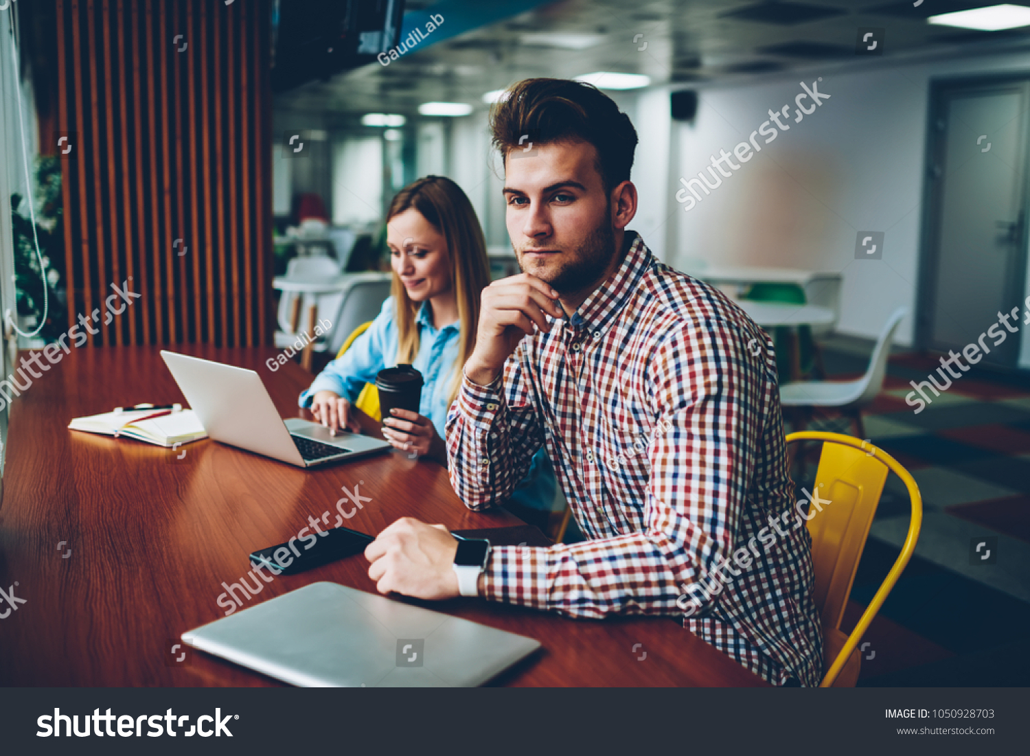 Thoughtful young man in casual shirt thinking on idea for coursework while hipster girl with tasty coffee searching information on websites on laptop computer connected to wireless internet #1050928703