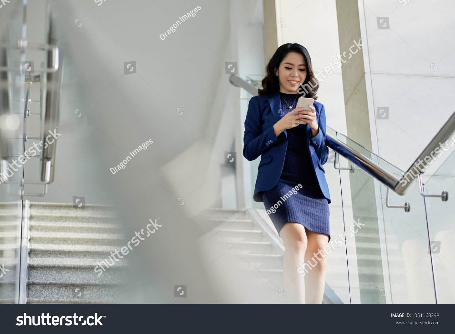 Smiling Asian manager in formalwear texting with friend on smartphone while going downstairs, portrait shot #1051168298
