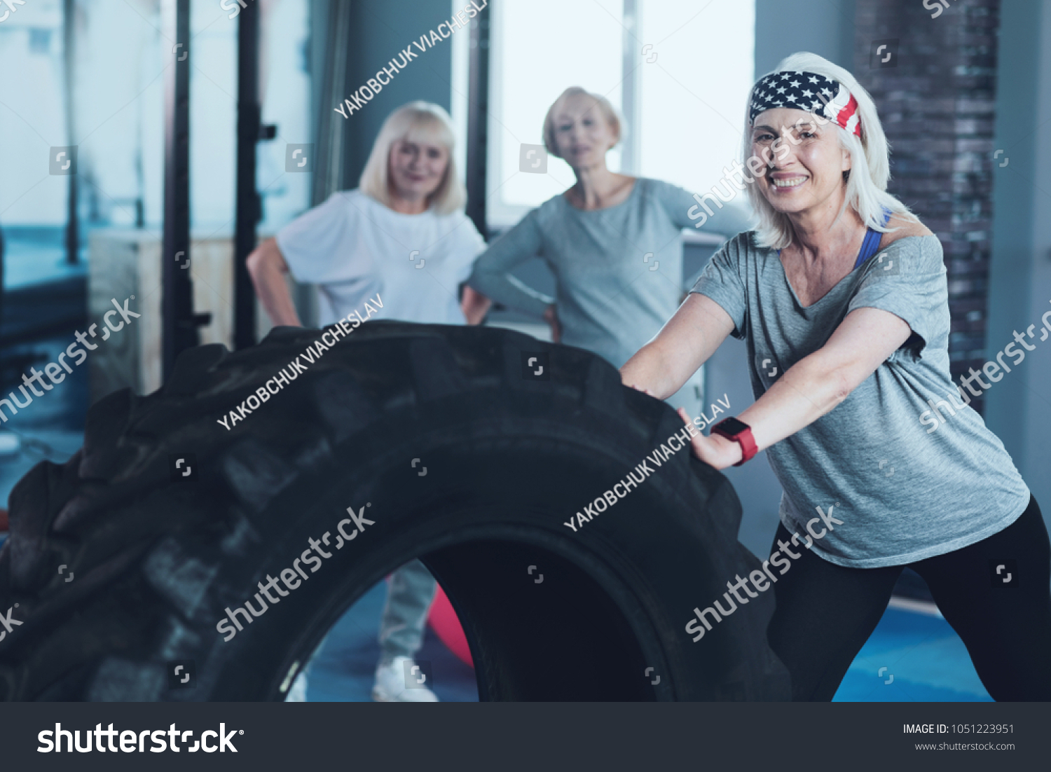 No more boundaries. Selective focus on a sporty retired lady looking into the camera with a smile on her face while training with a heavy wheel at a gym. #1051223951