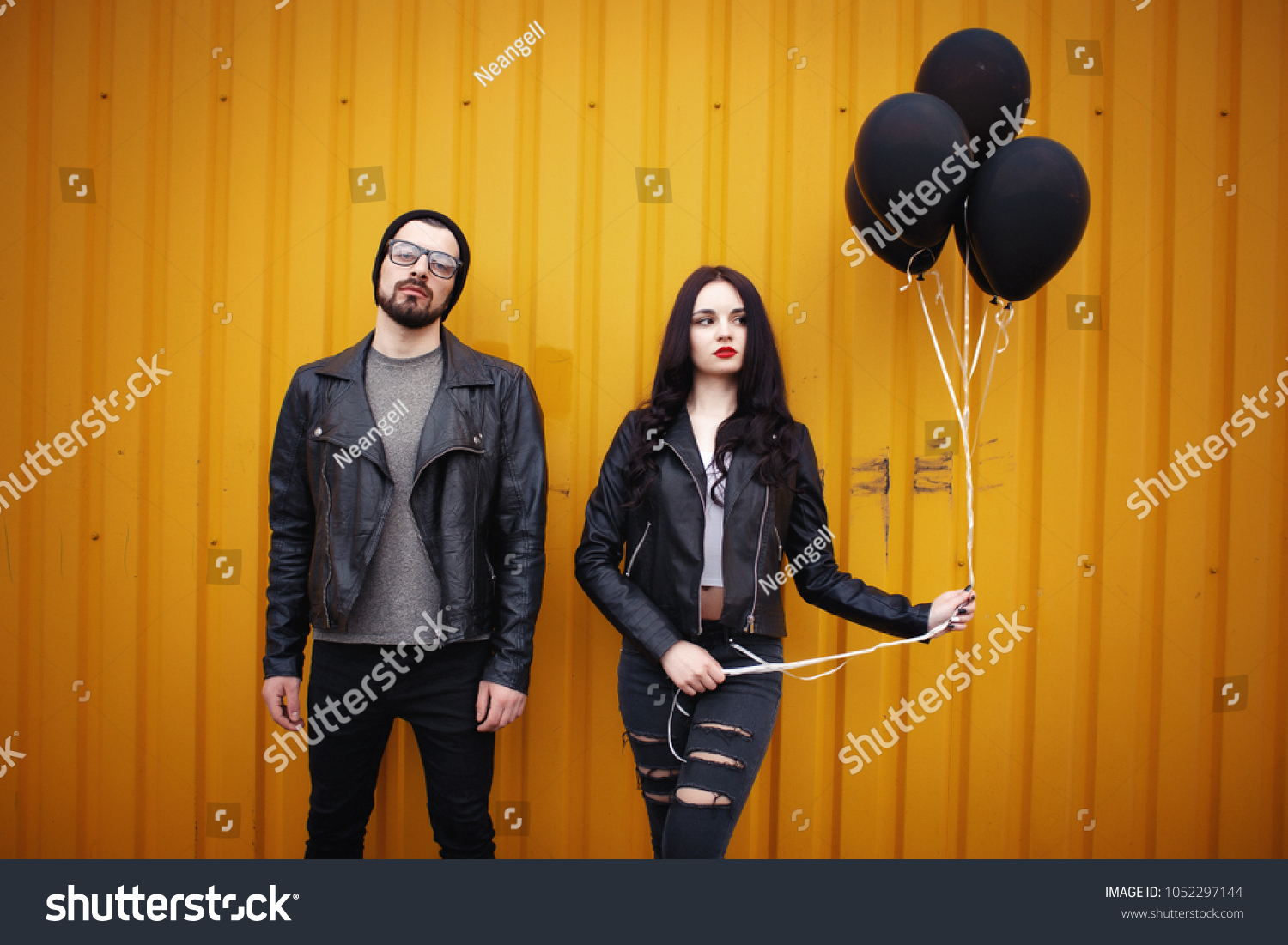 Stylish enamored guy and girl in leather jackets near a yellow green wall with black air balloons in hands #1052297144