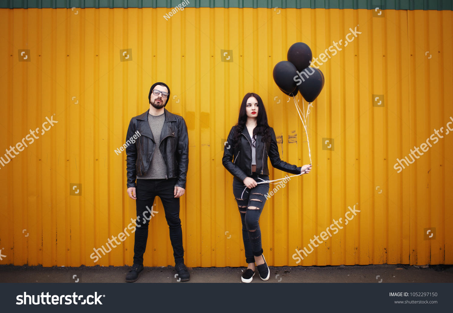 Stylish enamored guy and girl in leather jackets near a yellow green wall with black air balloons in hands #1052297150