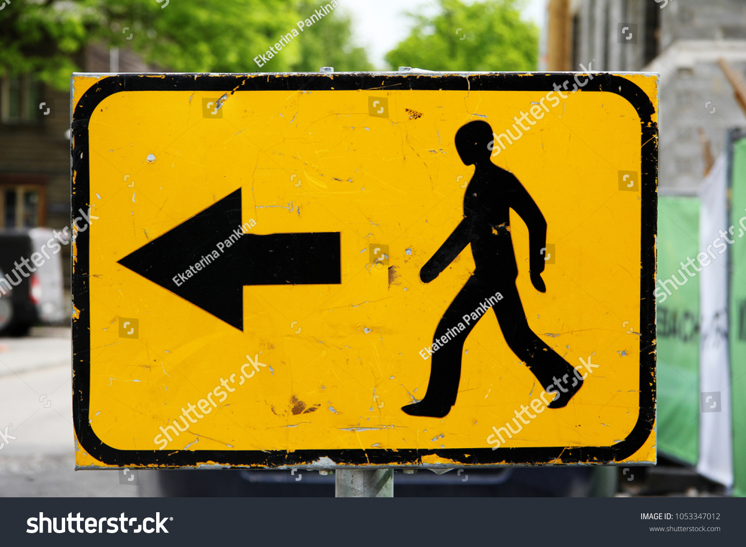 Traffic sign: on a yellow background a black silhouette of a walking man and a black arrow to the left against a city landscape background. Signs, symbols. #1053347012