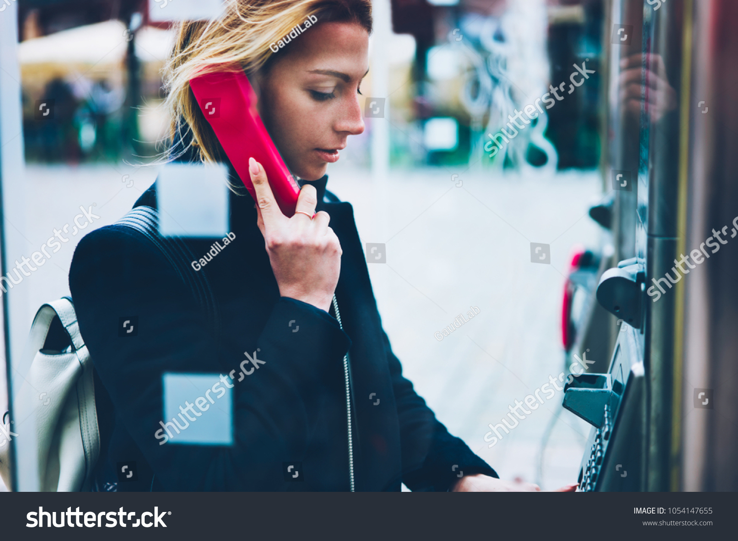 Thoughtful female tourist making call in payphone on street standing in transparent booth, young woman using public telephone operated with coins during travel for call with low prices abroad #1054147655