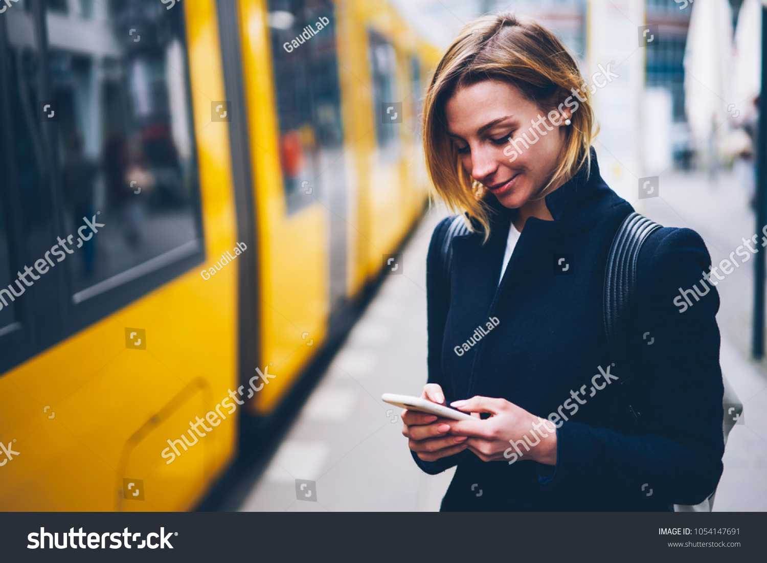 Positive blonde woman holding cellular updating information about city transport on web page, smiling hipster girl satisfied with online ticket service paying for electric transport via smartphone #1054147691