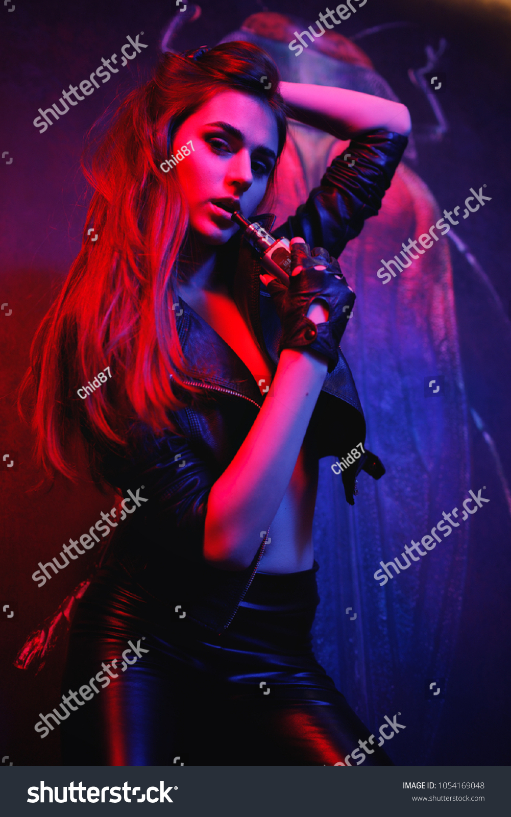 Young sexy woman is vaping ( smoking ) e-cigarette. Brutal sexy girl in black jacket and leather pant. Sexy rock star girl smoke electric cigarette in leather jacket, leather leggings and high heels. #1054169048