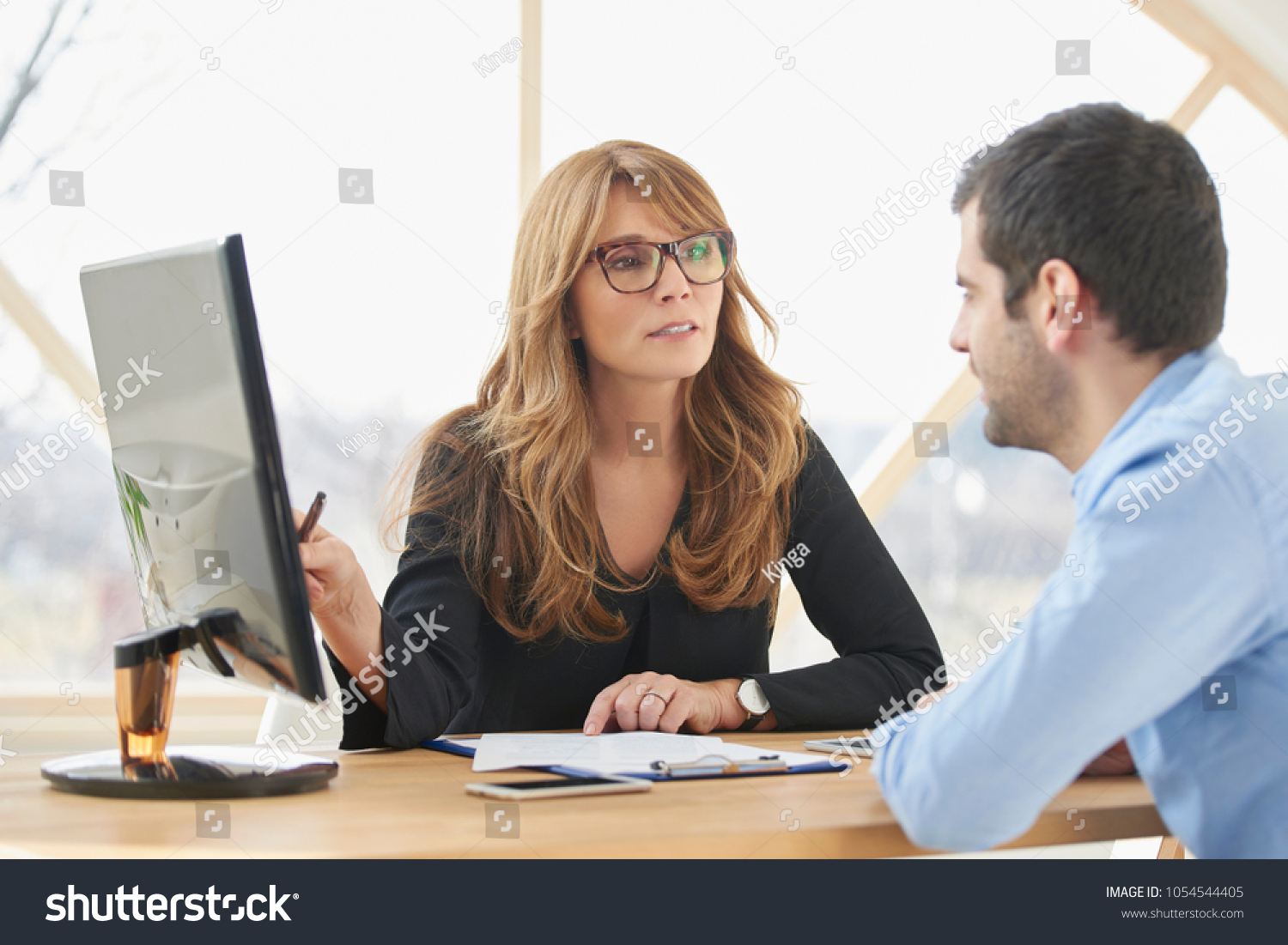 Executive middle aged businesswoman sitting at desk in front of laptop and doing some paperwork while giving sales advice to her young assistant. Brainstorming at the office. #1054544405