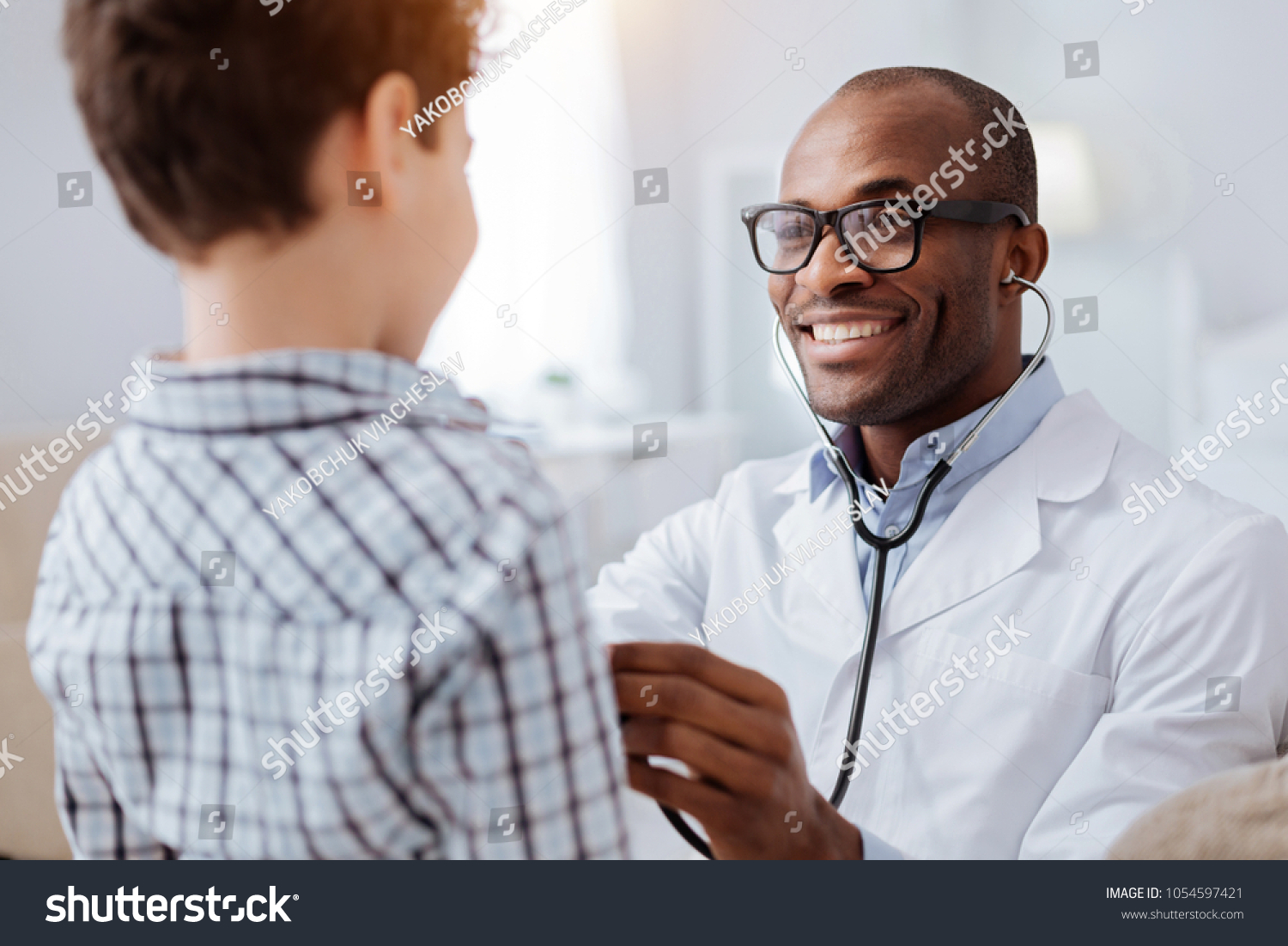 Stethoscope exam. Attractive cheerful male doctor listening to boy while putting on glasses and using stethoscope #1054597421