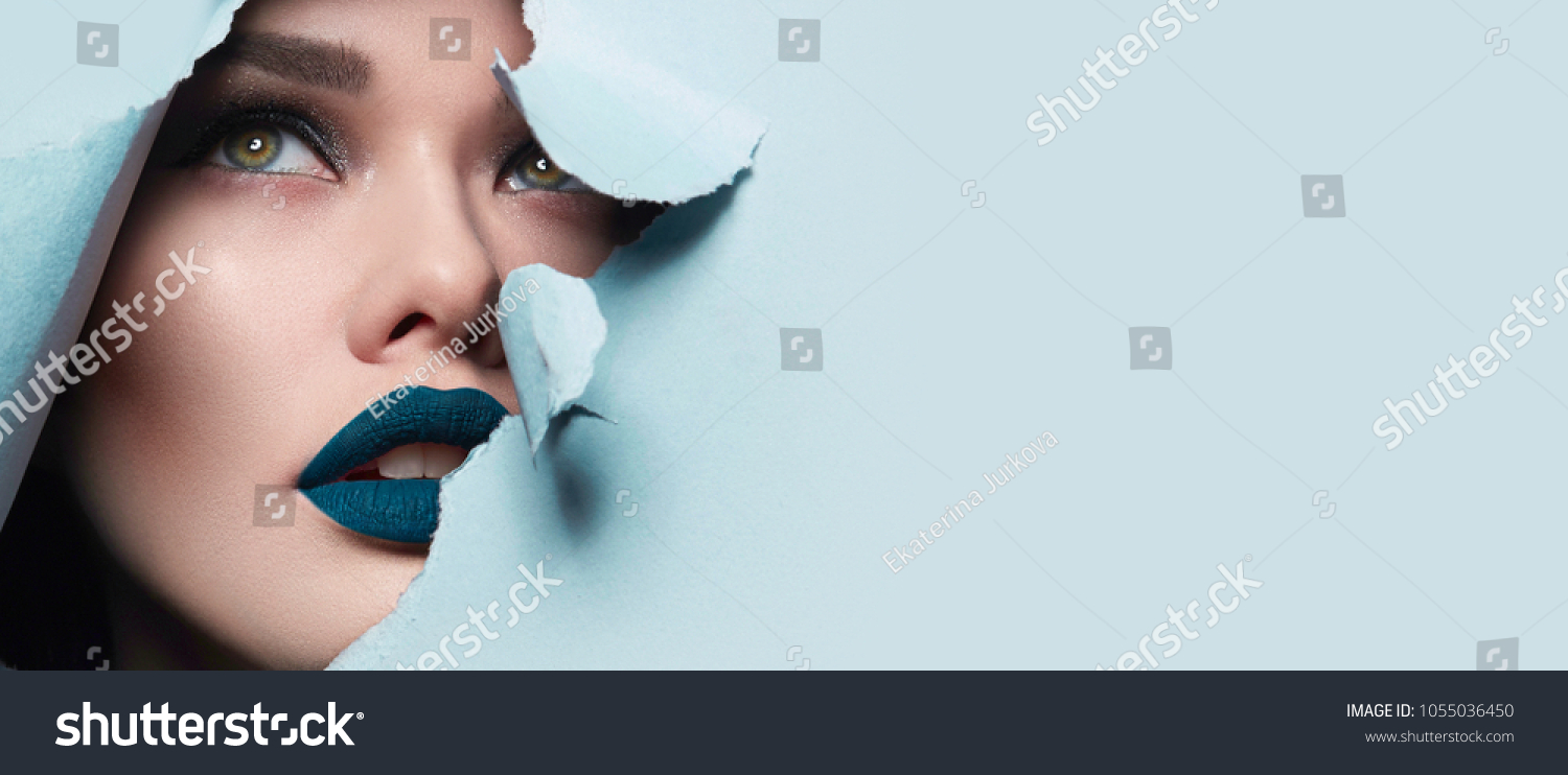 the face of a young beautiful girl with a bright make-up and puffy blue lips peers into a hole in blue paper.Fashion, beauty, make-up, cosmetics, hairstyle, beauty salon, boutique, discounts, sales. #1055036450
