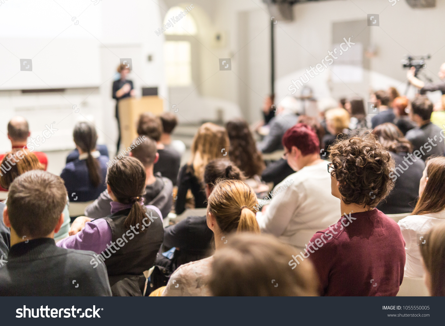 Business and entrepreneurship symposium. Female speaker giving a talk at business meeting. Audience in conference hall. Rear view of unrecognized participant in audience. Copy space on whitescreen. #1055550005