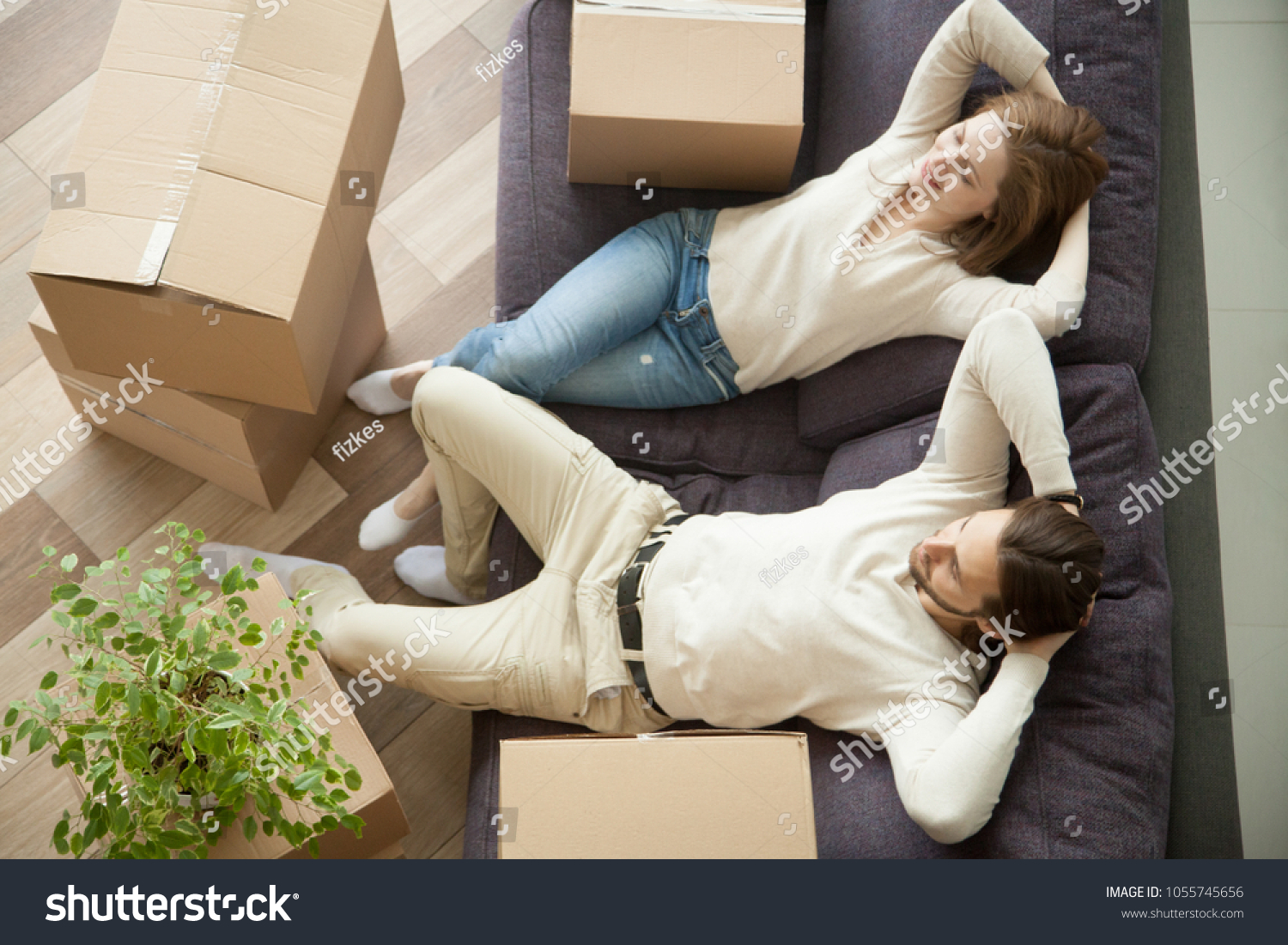 Relaxed smiling couple resting on couch moved in new home with boxes, happy real estate renters owners relaxing on sofa in living room, easy relocation with moving delivery service concept, top view #1055745656