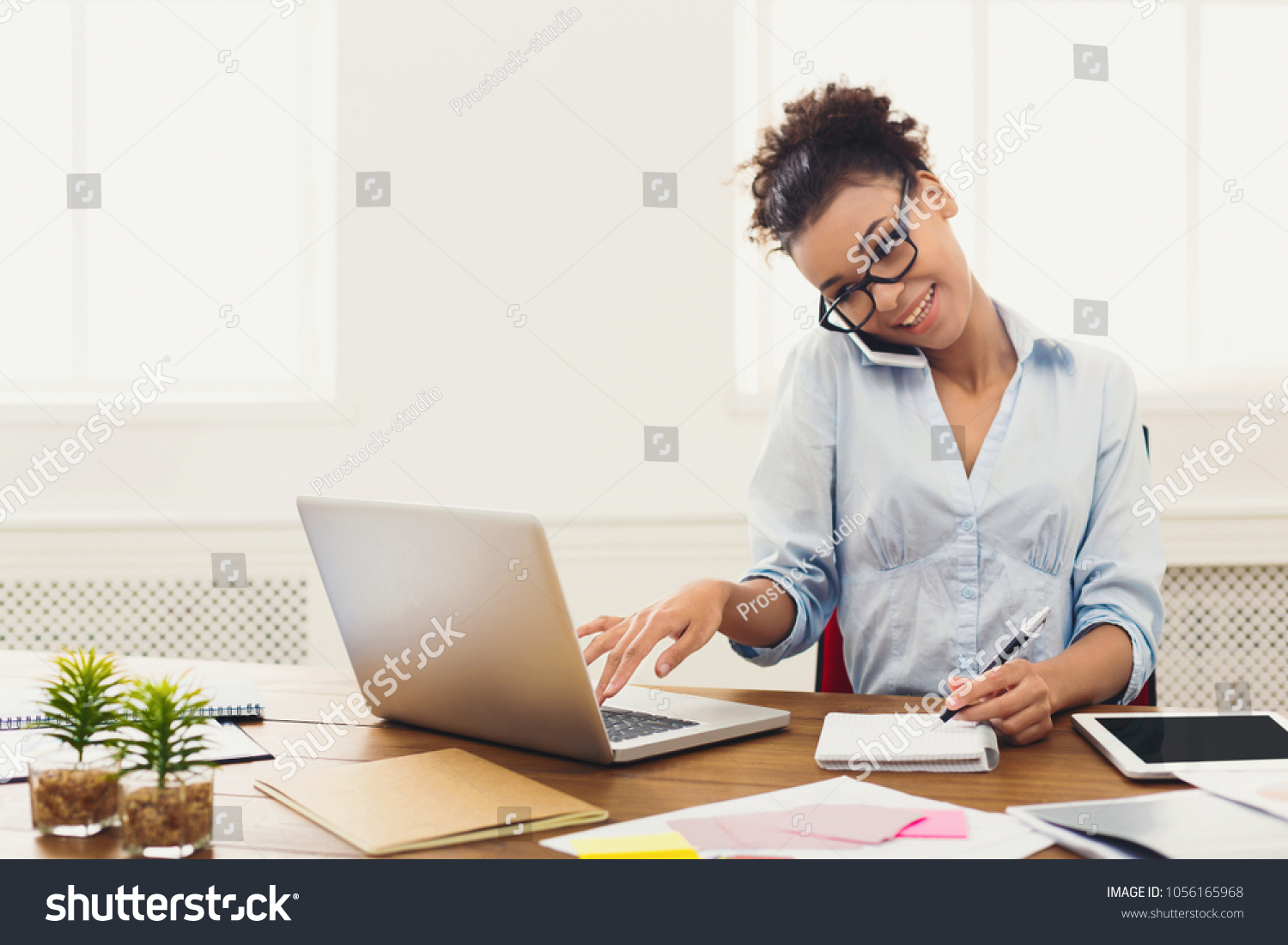 Happy african-american business woman talking on mobile at office. Young female manager consulting on phone, using laptop at working place, copy space #1056165968