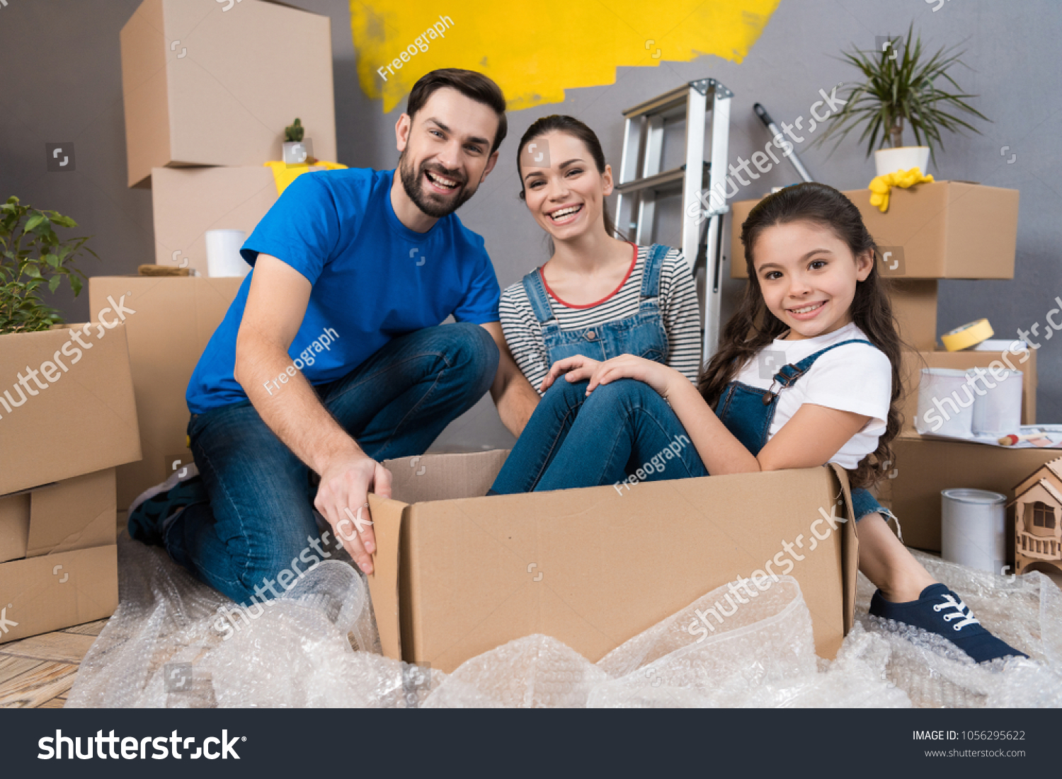 Home repair. Moving young family to new apartment. Repair in house for sale. Happy couple carries small daughter in box at house where repairs are being carried out. #1056295622