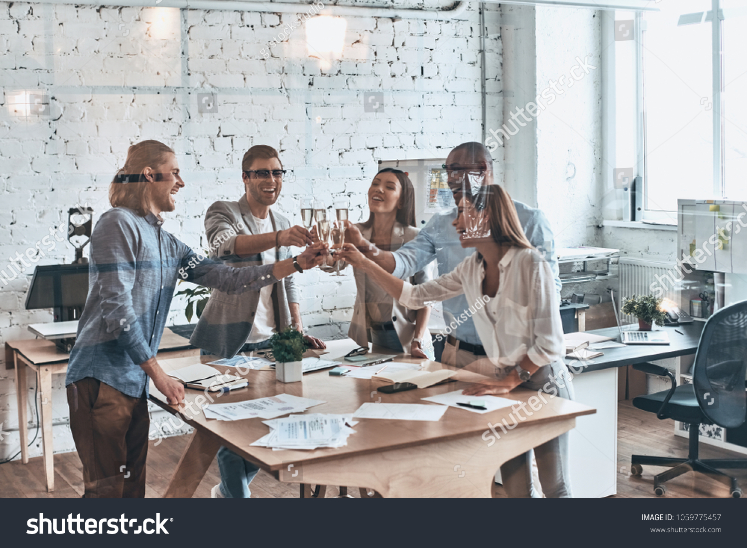 Toasting their success. Group of young business people toasting each other and smiling while standing behind the glass wall in the board room #1059775457