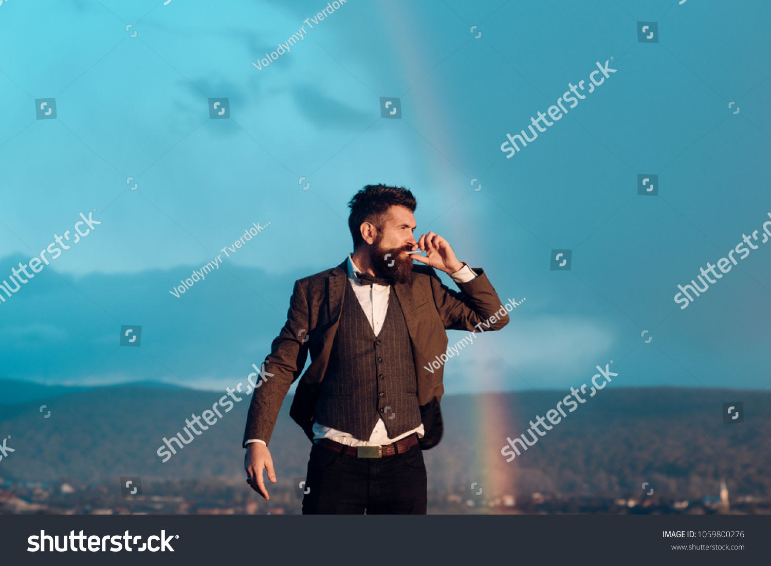 Hipster with stylish appearance smoking in front of sky with rainbow. Lord of world concept. Successful man with scenery on background. Guy with strict face in suit feels free and successful. #1059800276