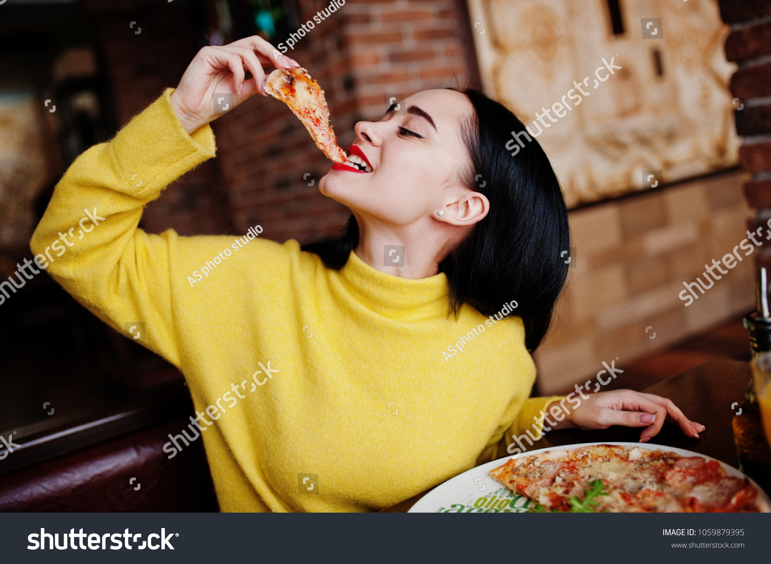 Funny brunette girl in yellow sweater eating pizza at restaurant.  #1059879395