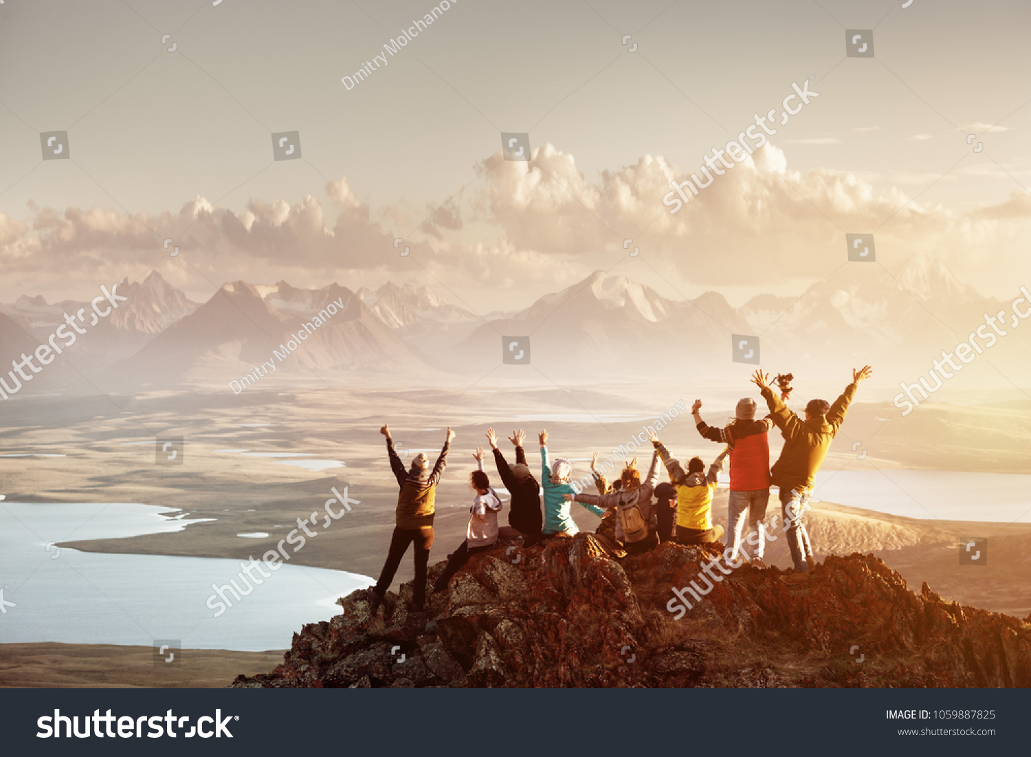Big group of people having fun in success pose with raised arms on mountain top against sunset lakes and mountains. Travel, adventure or expedition concept