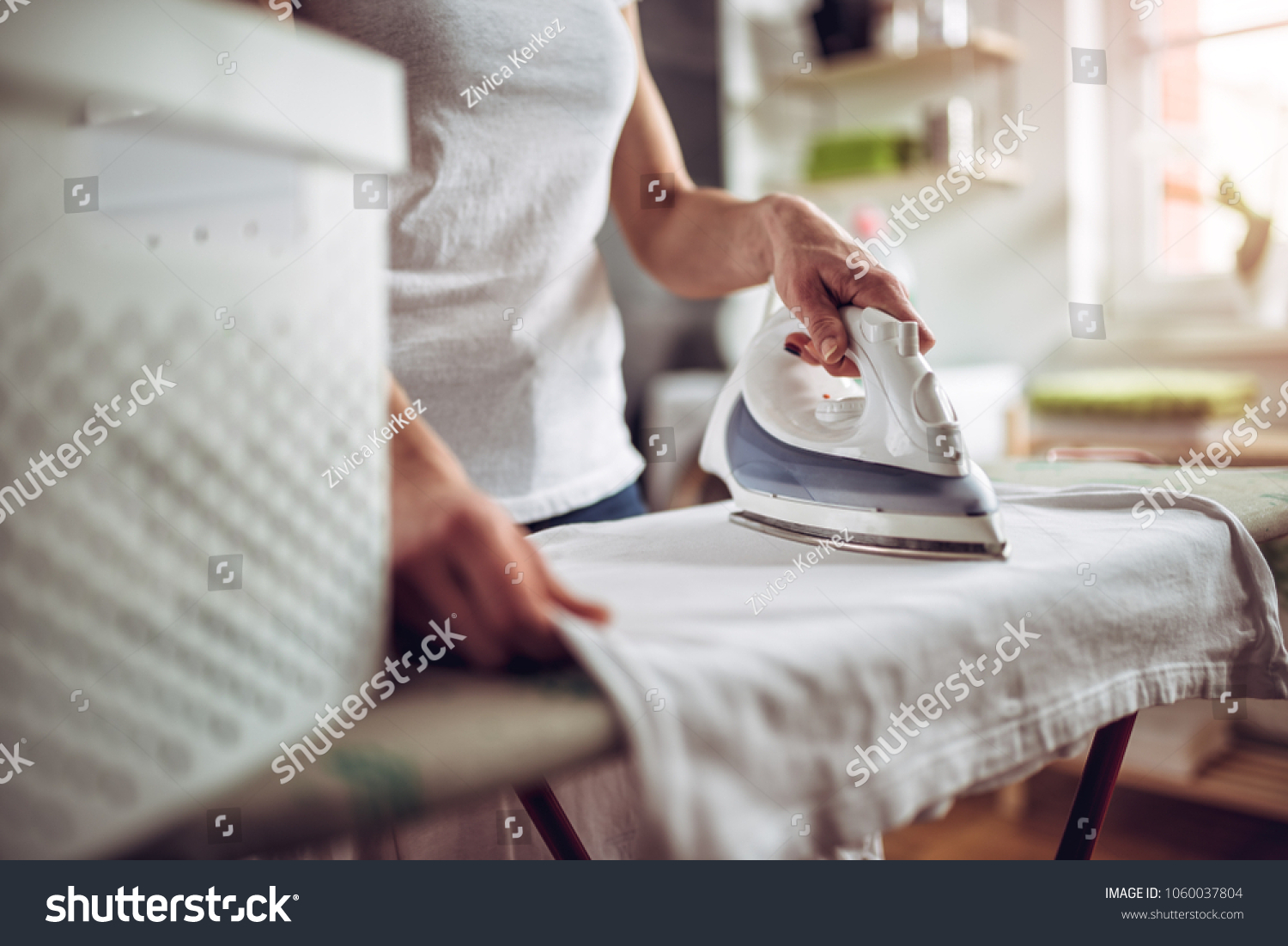 Women wearing white shirt ironing clothes on ironing board in laundry room at home #1060037804