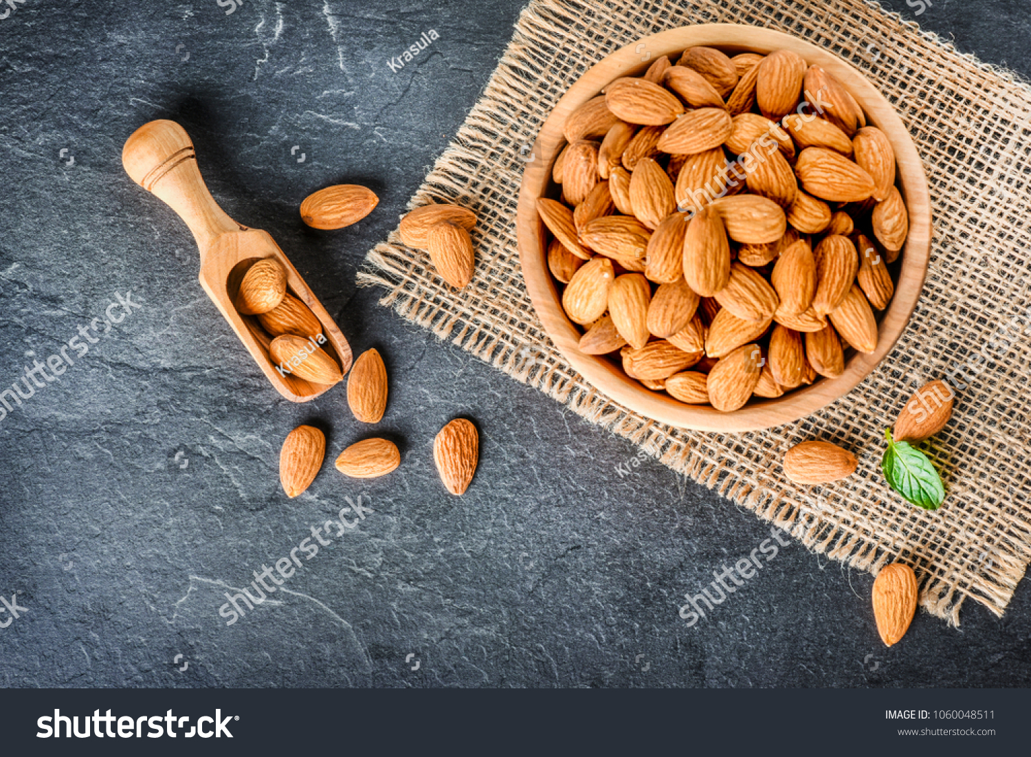 Top view of almonds on dark stone table with wood spoon or scoop. Almond in wooden bowl. Nuts freely laid on dark board. #1060048511