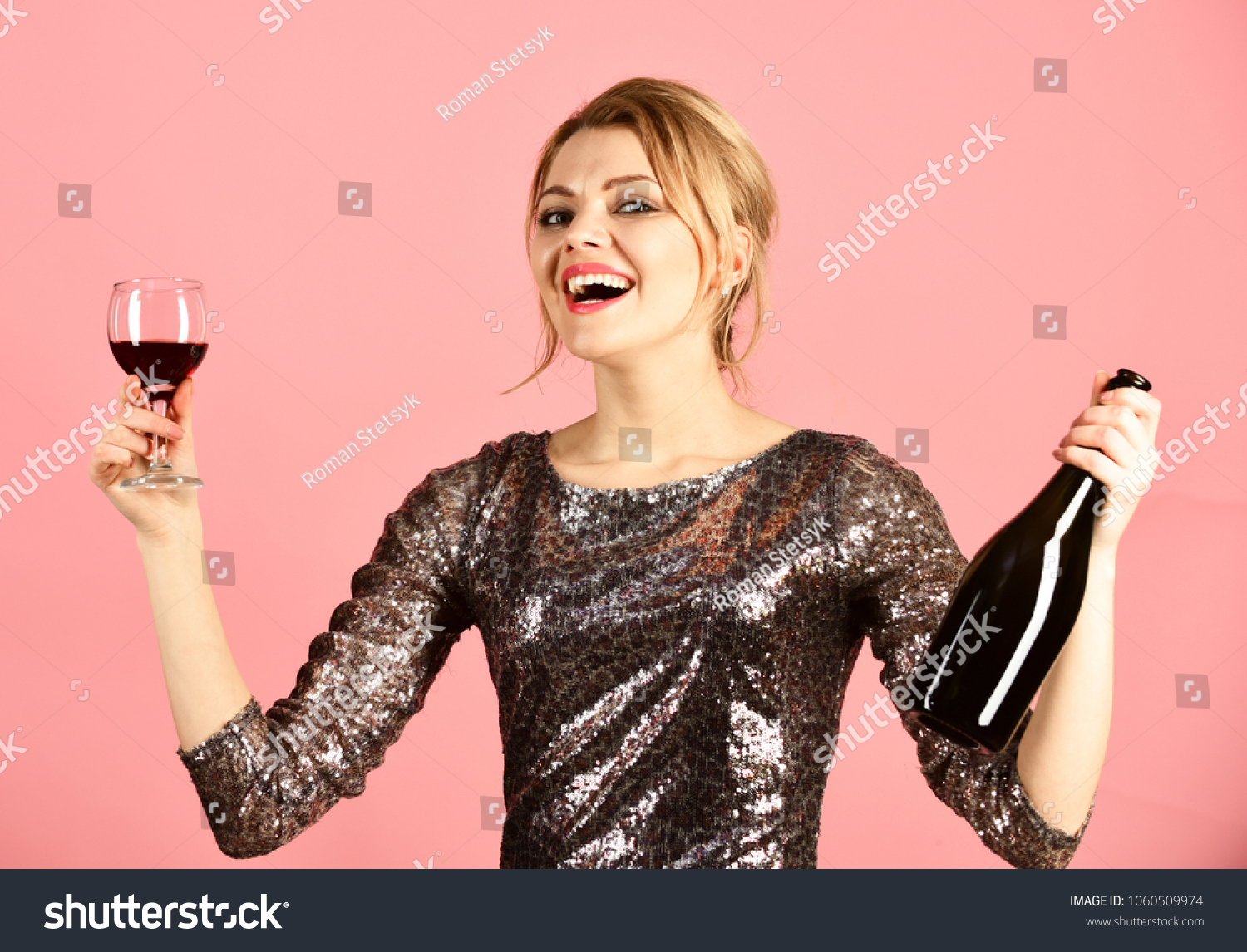 Girl in shining dress with alcohol on pink background. Lady holding glass and bottle of red Italian wine. Winetasting or party concept. Woman with smiling face drinks expensive cabernet or merlot. #1060509974