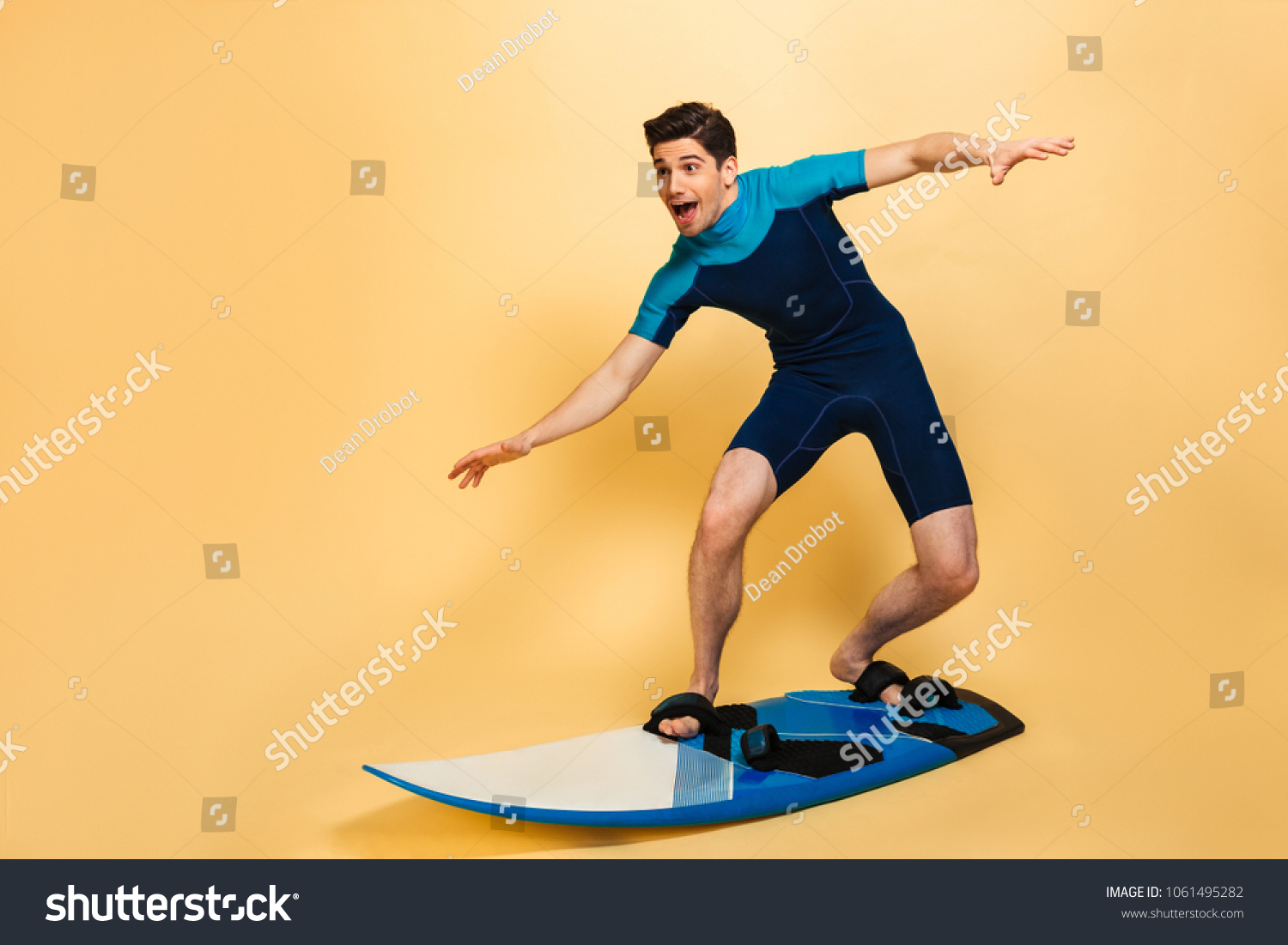 Full length portrait of an excited young man dressed in swimsuit surfing on a board isolated over yellow background #1061495282