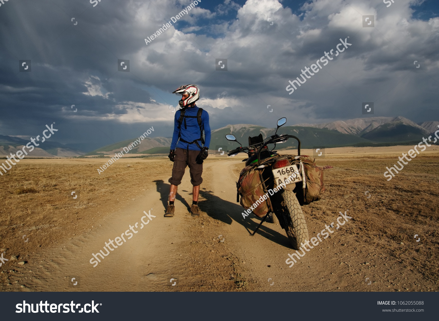 Motorcycle traveler man in helmet with suitcases standing on extreme rocky road in a mountain valley in cloudy weather on the background of endless steppe #1062055088