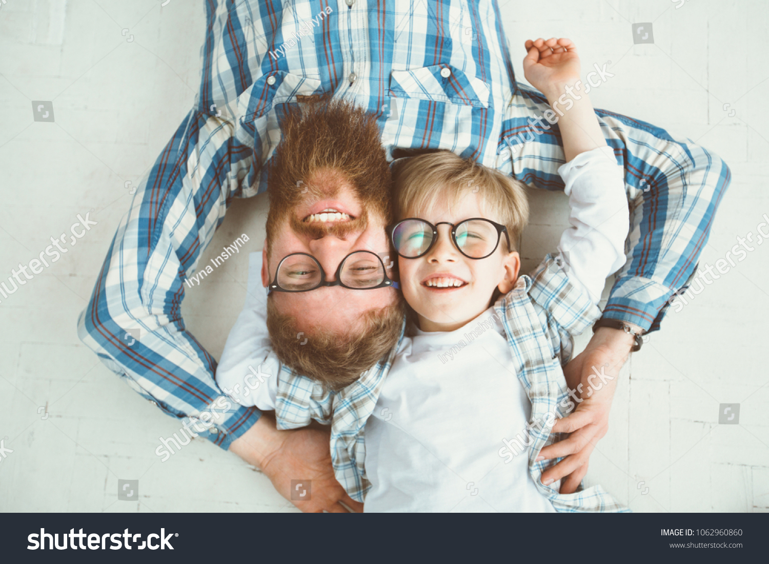 Top view of cute little boy and his handsome young beard dad, both in eyeglasses, smiling while lying with hands behind head on white floor. #1062960860