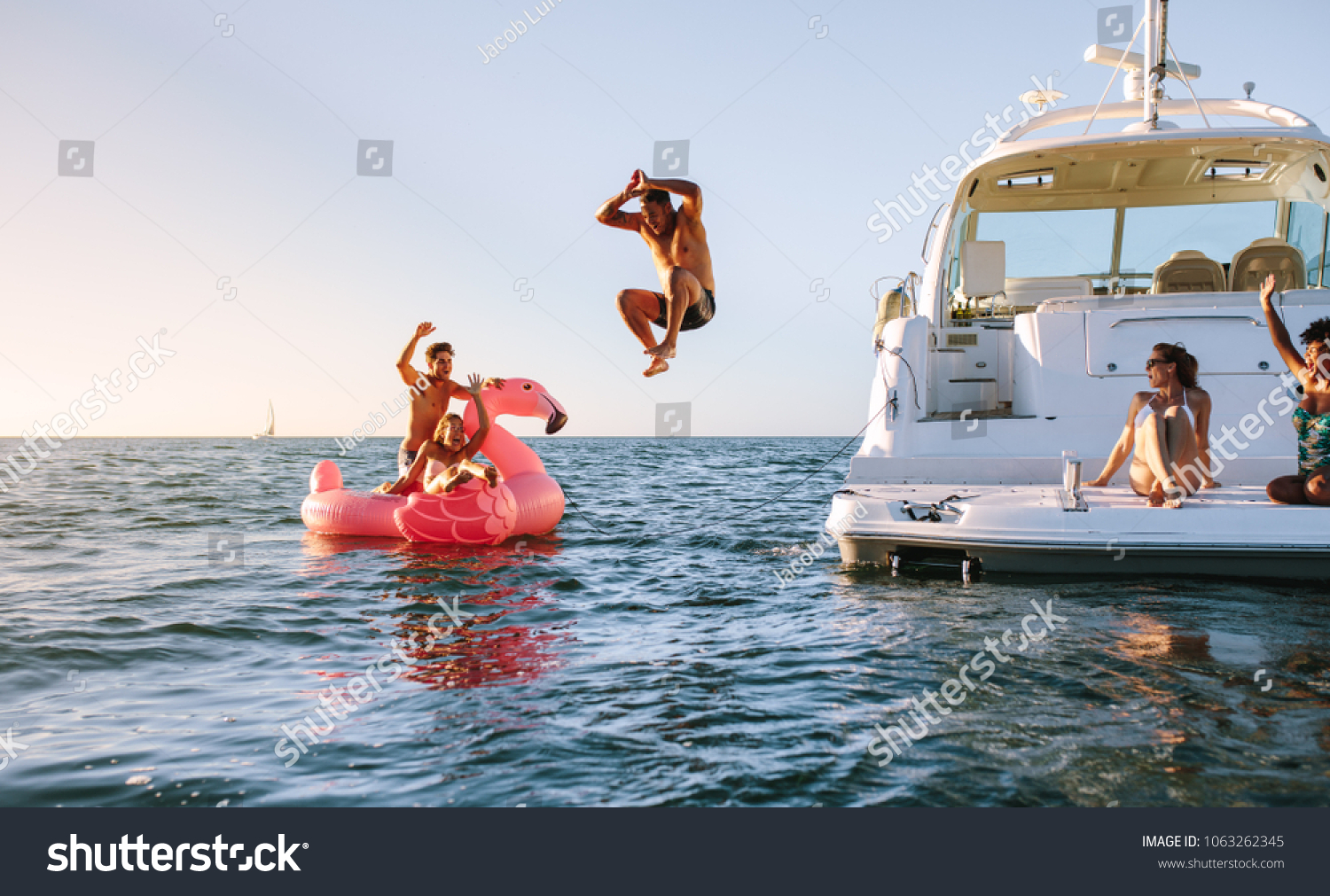 Man diving in the sea with friends sitting on yacht and inflatable toy. Group of friends enjoying a summer day on a inflatable toy and yacht. #1063262345
