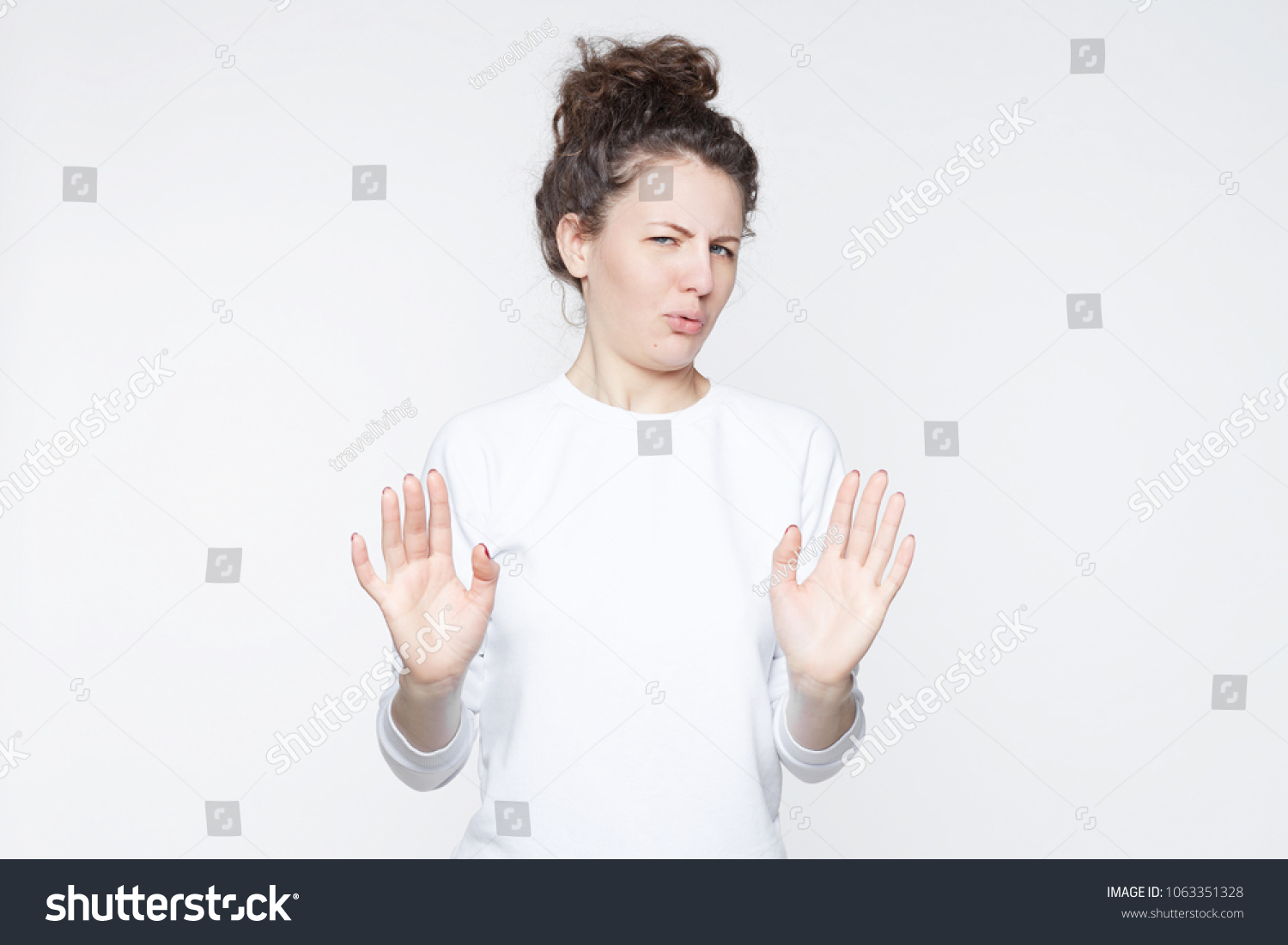 Body language. Disgusted stressed out beautiful young woman with hair knot posing at studio wall, keeping hands in stop gesture, grimacing, trying to defend herself as if saying: Stay away from me. #1063351328