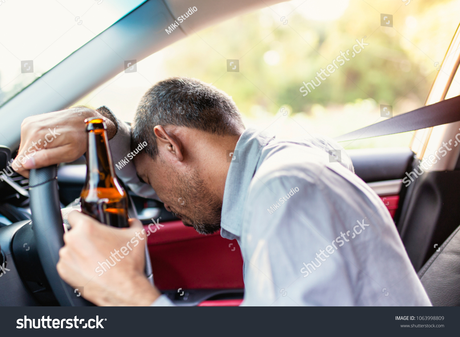 Drunk asian young man drives a car with a bottle of beer with sunset background, Dangerous driving concept #1063998809