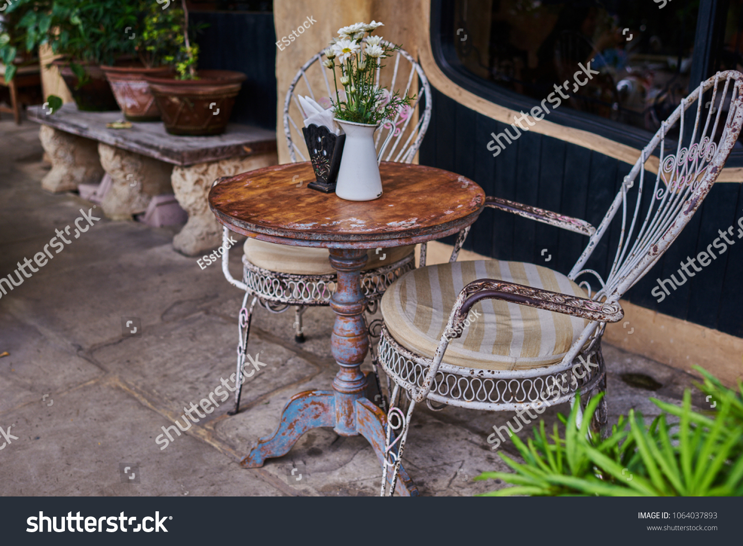 Old white antique wrought iron furniture, shabby chic exterior. Set of round table with flowers and two elegant chairs in the street in the old town. Table on cafe background. Vintage style.   #1064037893