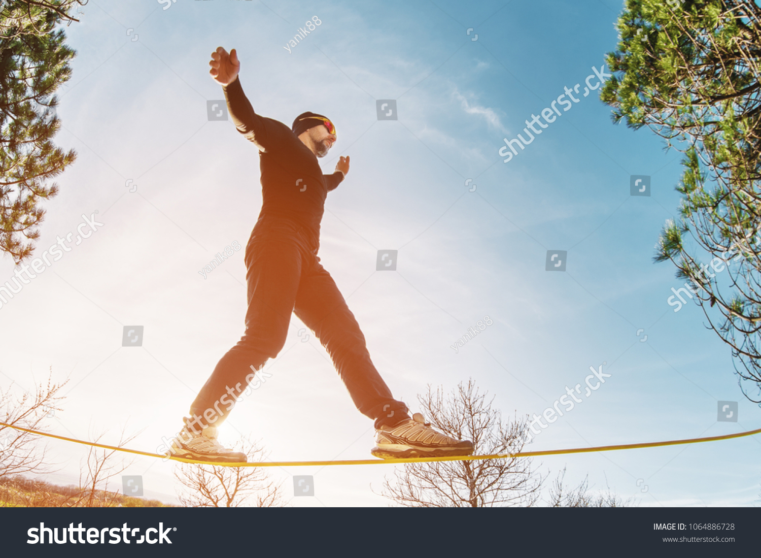 A man, aged with a beard and wearing sunglasses, balances on a slackline in the open air between two trees at sunset #1064886728