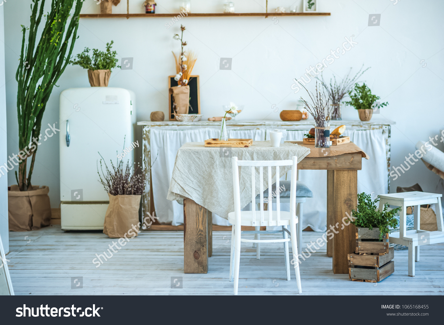 Beautiful spring photo of kitchen interior in light textured colors. Kitchen, living room with beige sofa sofa, old retro white fridge, rustic table, large cactus and woven macrame on the wall #1065168455