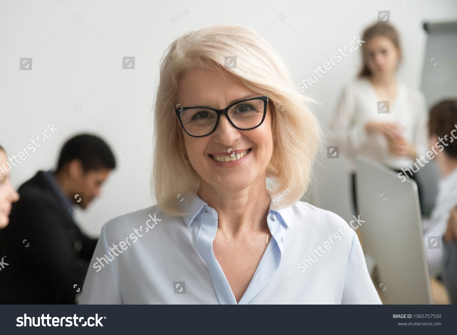 Portrait of smiling senior businesswoman wearing glasses with businesspeople at background, happy older team leader, female aged teacher professor or executive woman boss looking at camera, head shot #1065757550