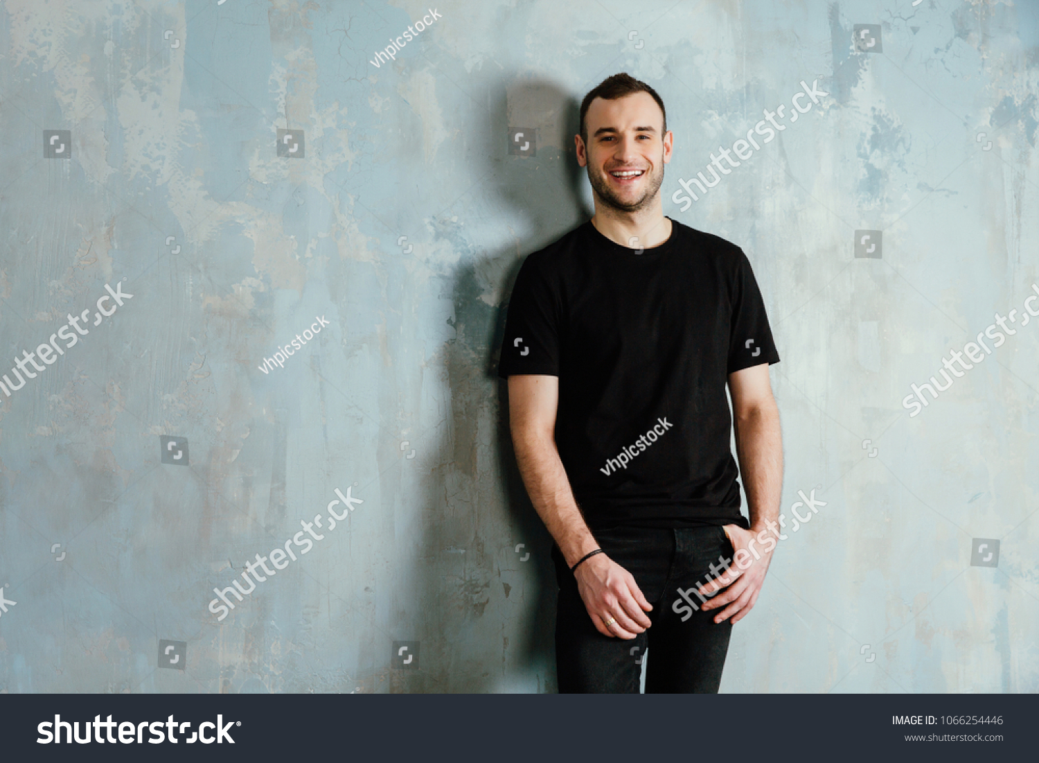 portrait of a young man in a black T-shirt leaned against a vintage gray wall. Copy space. Masculine model of bristles. Happy smiling. #1066254446