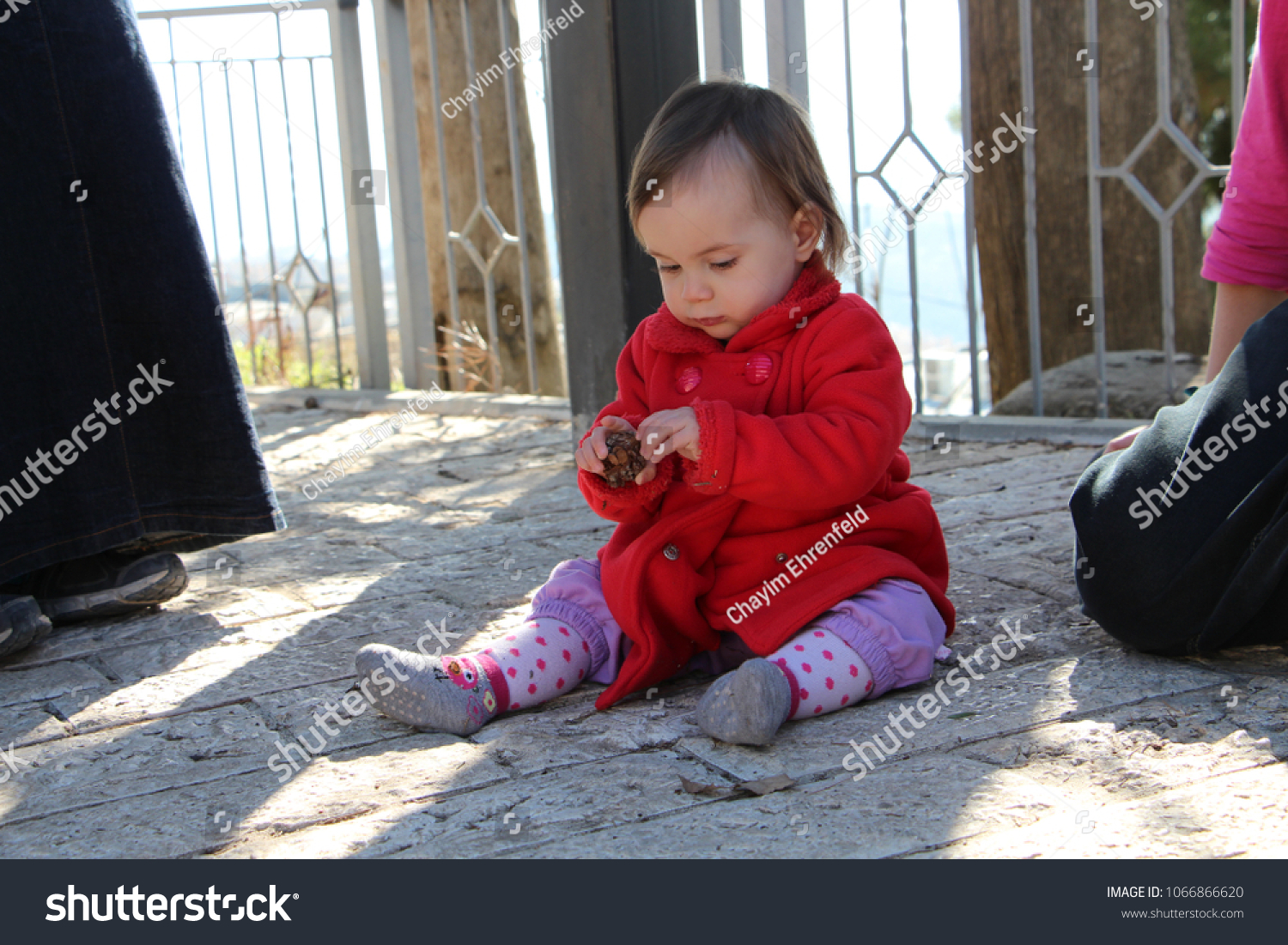Baby girl in red coat sitting on the sidewalk, playing with a pinecone
 #1066866620