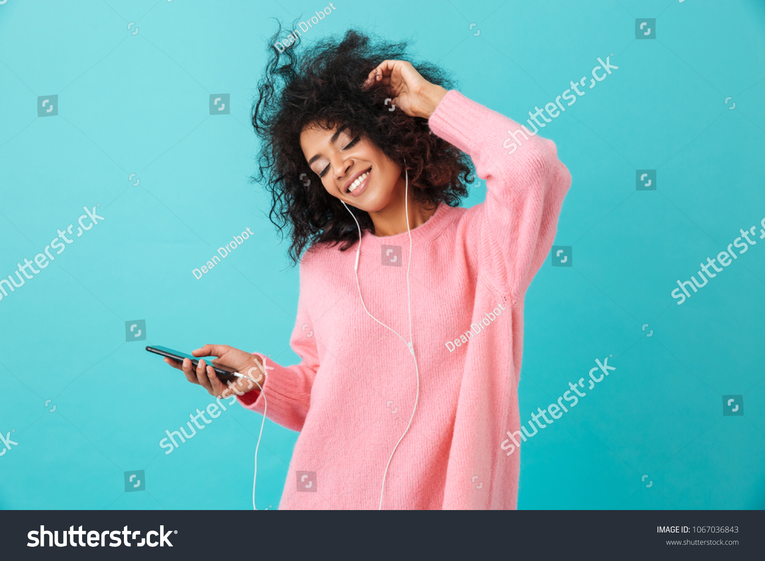 Joyous american woman in casual clothing dancing and listening to music with pleasure via white earphones isolated over blue background #1067036843