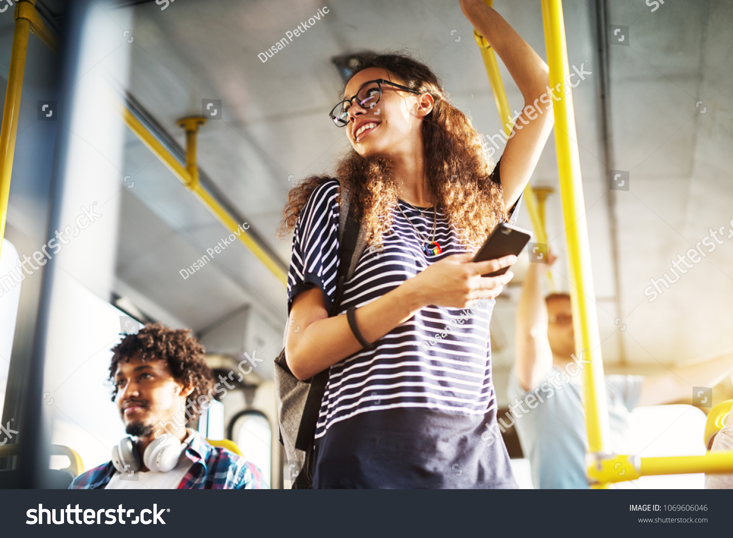 Young adorable joyful woman is standing on the bus using the phone and smiling. #1069606046